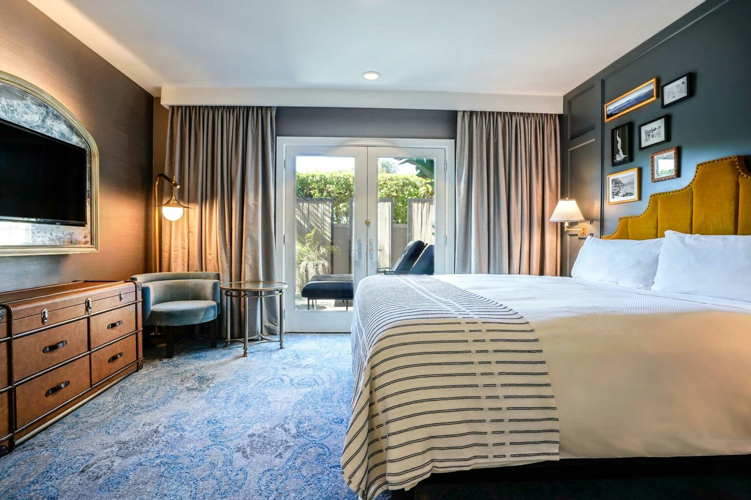 Stylish hotel room with a king-sized bed, mustard-yellow headboard, blue carpeting, and a cozy reading chair. Light streams through glass doors.