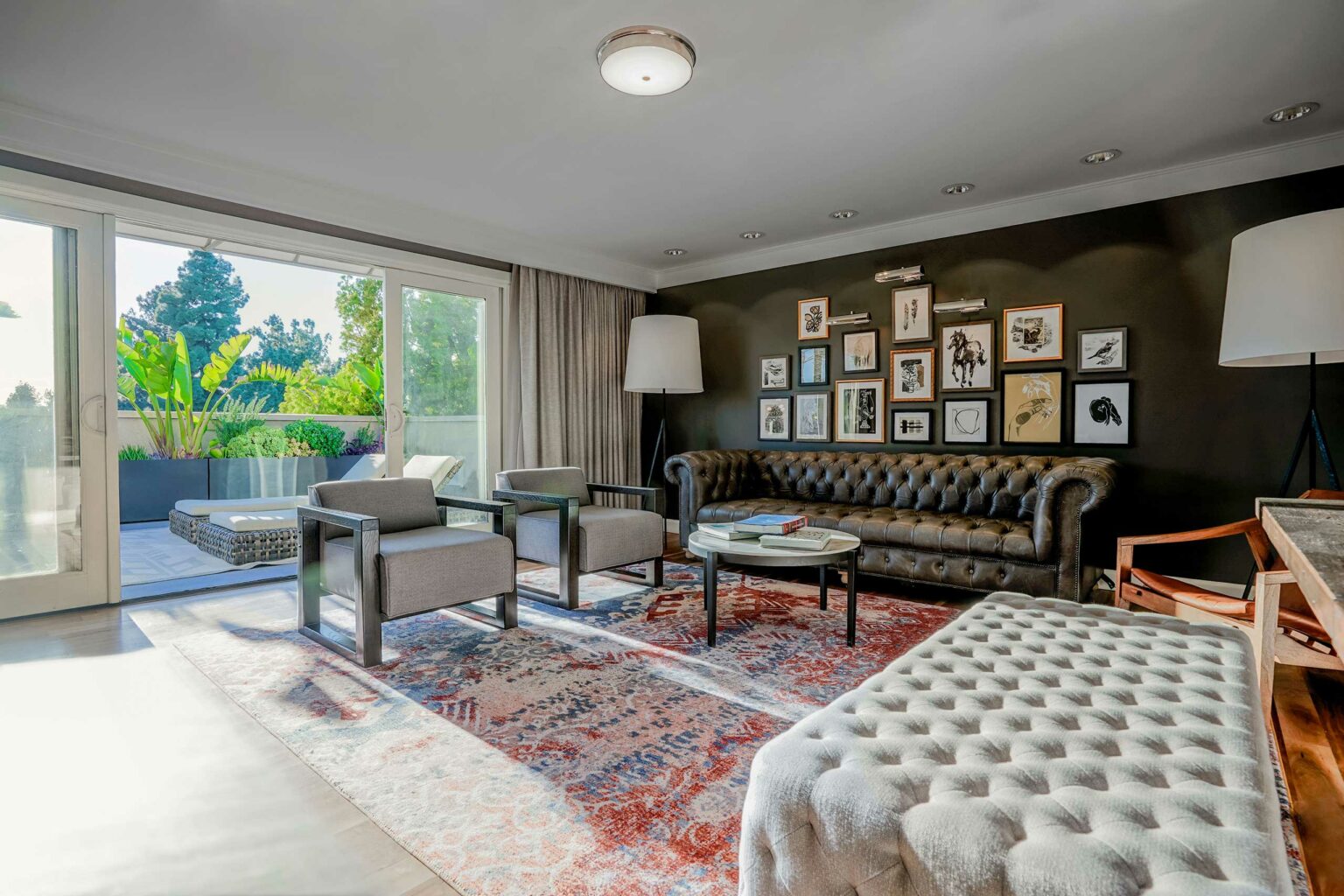 Chic guest room living room with a leather sofa, two armchairs, a patterned rug, and a gallery wall. Sunlight streams through large glass doors, showing a lush garden.