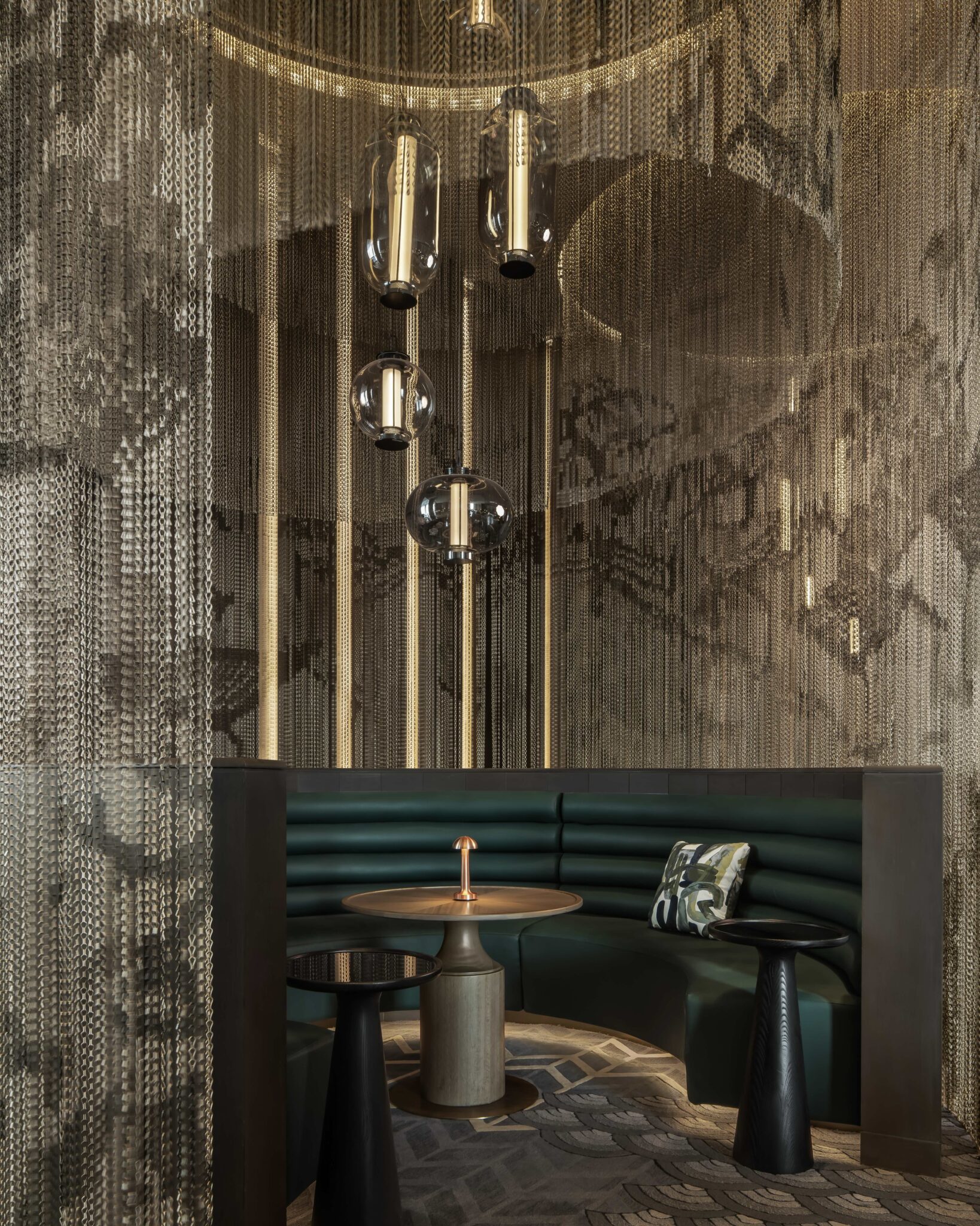 Luxurious hotel seating area with a curved green leather booth, modern hanging lights, and metallic chain curtains. Warm, elegant ambiance with art deco accents.