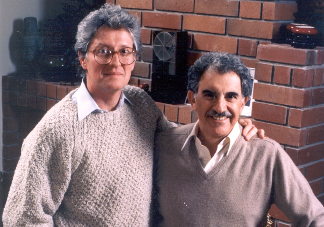 Michael Bedner and Howard Hirsch pose closely, smiling warmly. On the left, Michael Bedner wears a knitted sweater; on the right, Howard Hirsch wears a collared shirt and sweater vest. A brick wall is in the background, creating a cozy atmosphere.