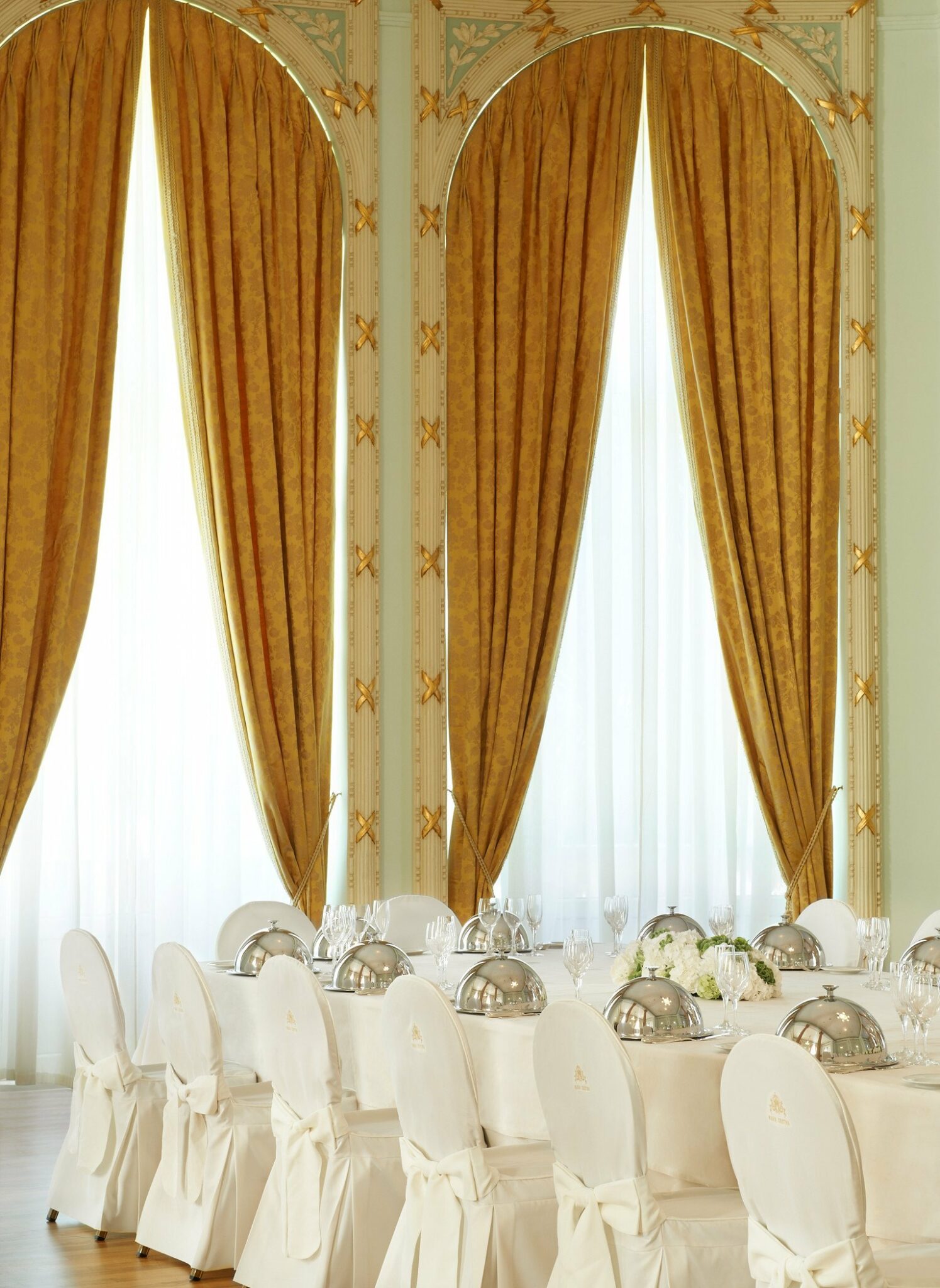 Elegant ballroom with tall, golden curtains and a set table for eight. White chairs surround the table, adorned with cloches and glassware. Luxurious ambiance.