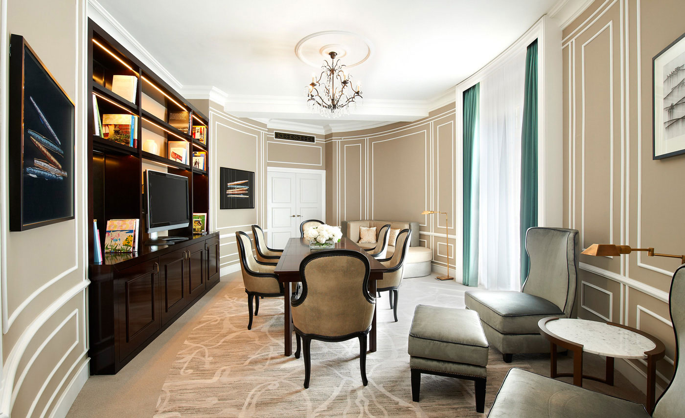 Elegant suite study room with beige walls, a large wooden table, plush chairs, a chandelier, and a built-in shelf with books. Soft, calming atmosphere.