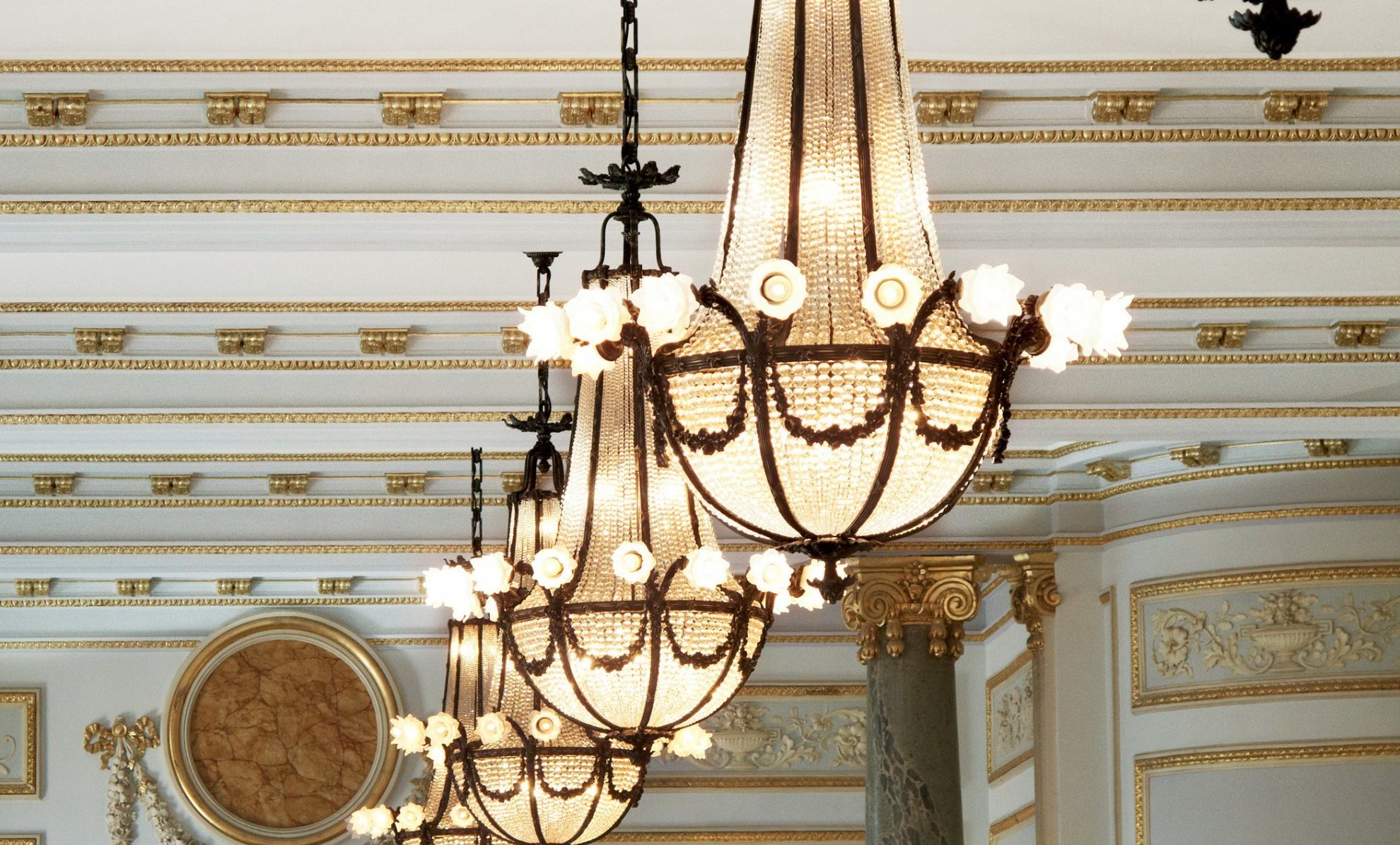 Elegant chandeliers with crystal details hang from an ornate ceiling with gold accents and intricate molding, exuding a luxurious ambiance.