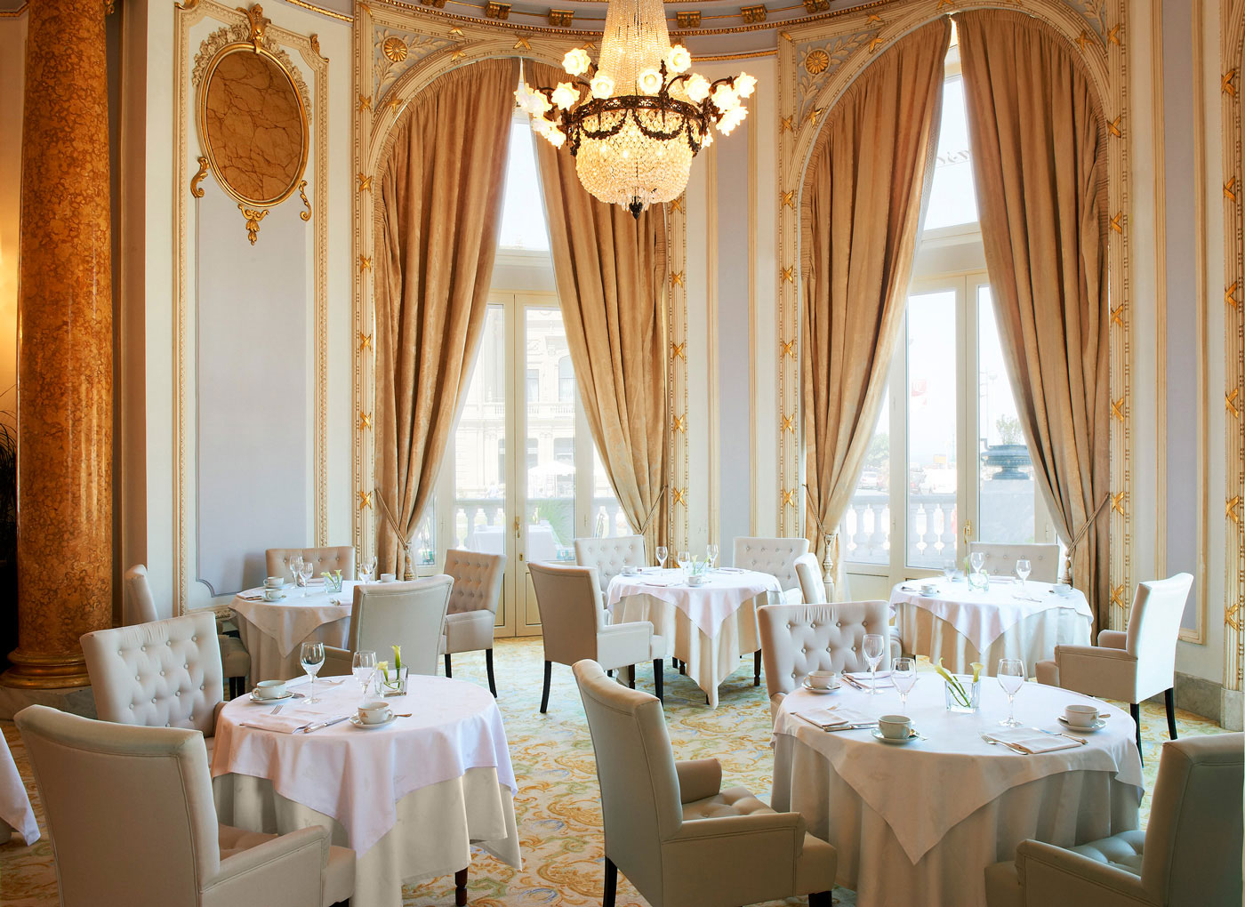 Luxurious dining room with elegant decor, featuring round tables with white tablecloths, plush beige chairs, large draped windows, and a grand chandelier.