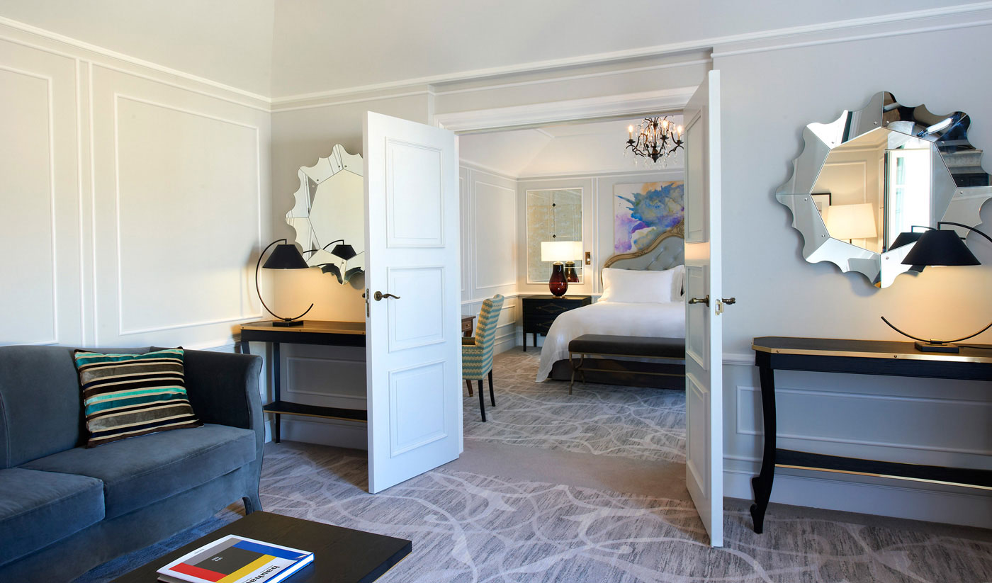 Bright, elegant suite living room with a plush gray sofa, decorative mirrors, and an inviting bedroom. Features modern decor, stylish lighting, and serene ambiance.