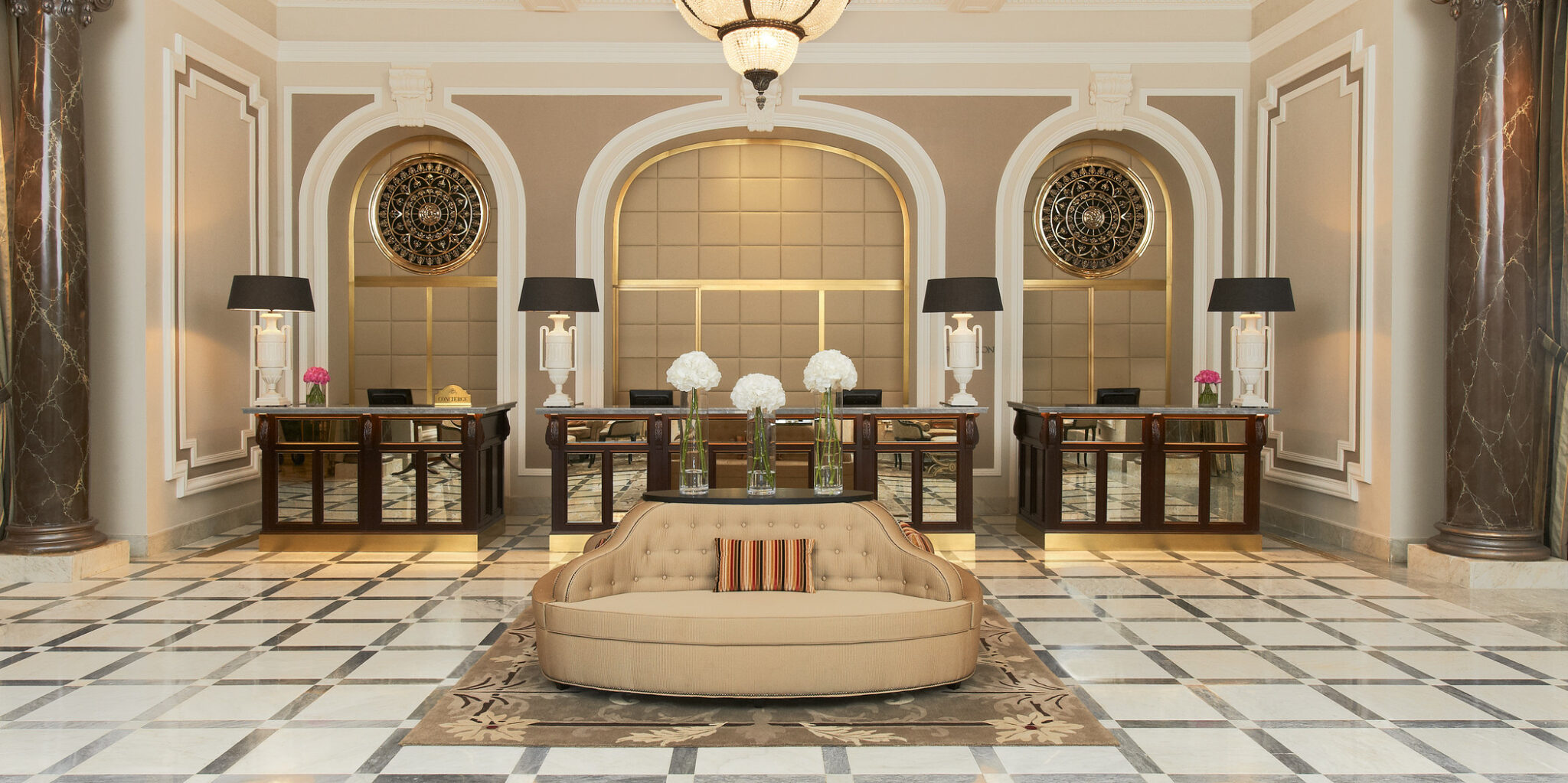 Elegant hotel lobby with beige couches, striped cushions, and glass vases filled with white flowers. Behind are desks with black lamps and decorative art.
