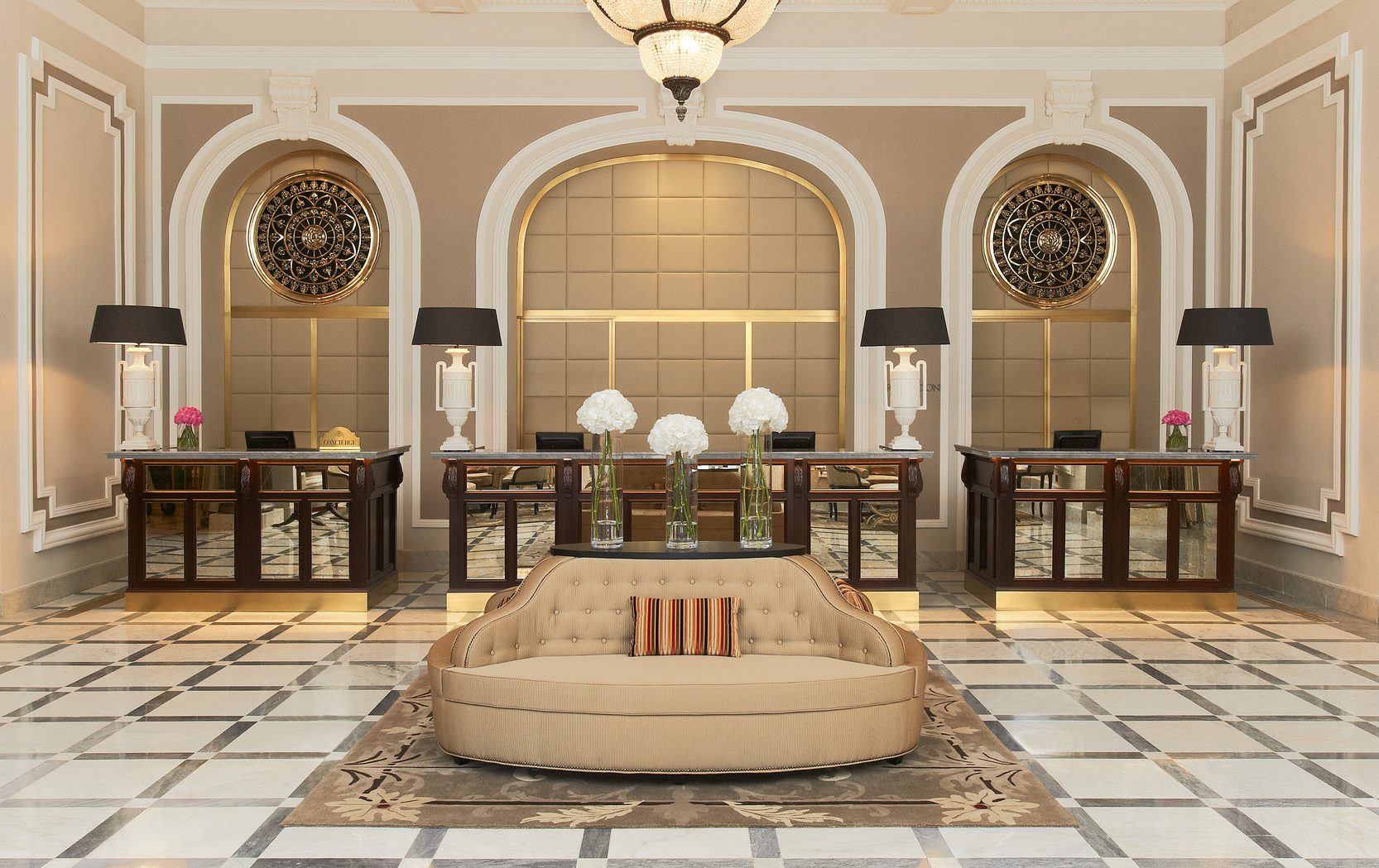 Elegant hotel lobby with beige couches, striped cushions, and glass vases filled with white flowers. Behind are desks with black lamps and decorative art.