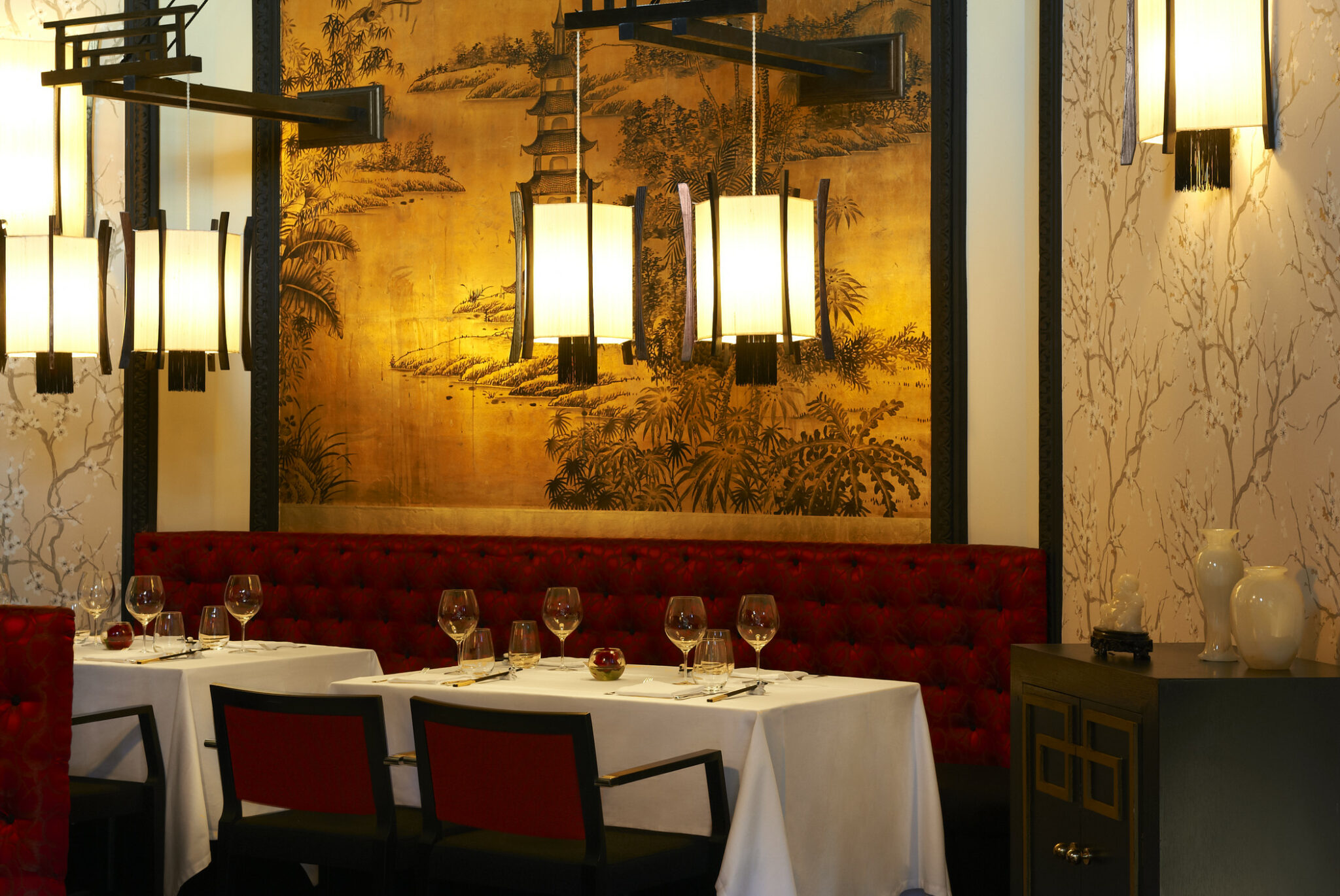 Elegant restaurant interior with two white-clothed tables, surrounded by red-padded chairs. A golden Asian-themed wall painting and warm pendant lights evoke a cozy, sophisticated atmosphere.