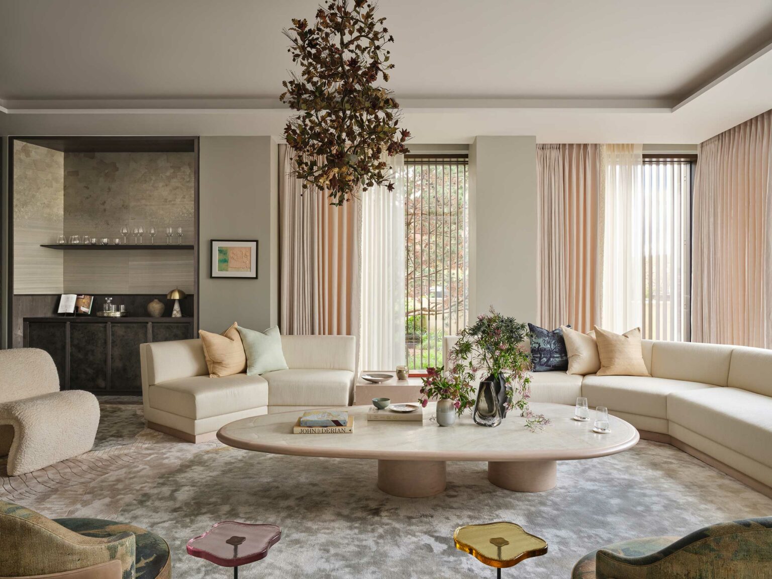 A cozy living room with beige sectional sofas, a large oval coffee table, floral centerpiece, and hanging leafy chandelier. Soft natural light filters through sheer curtains.