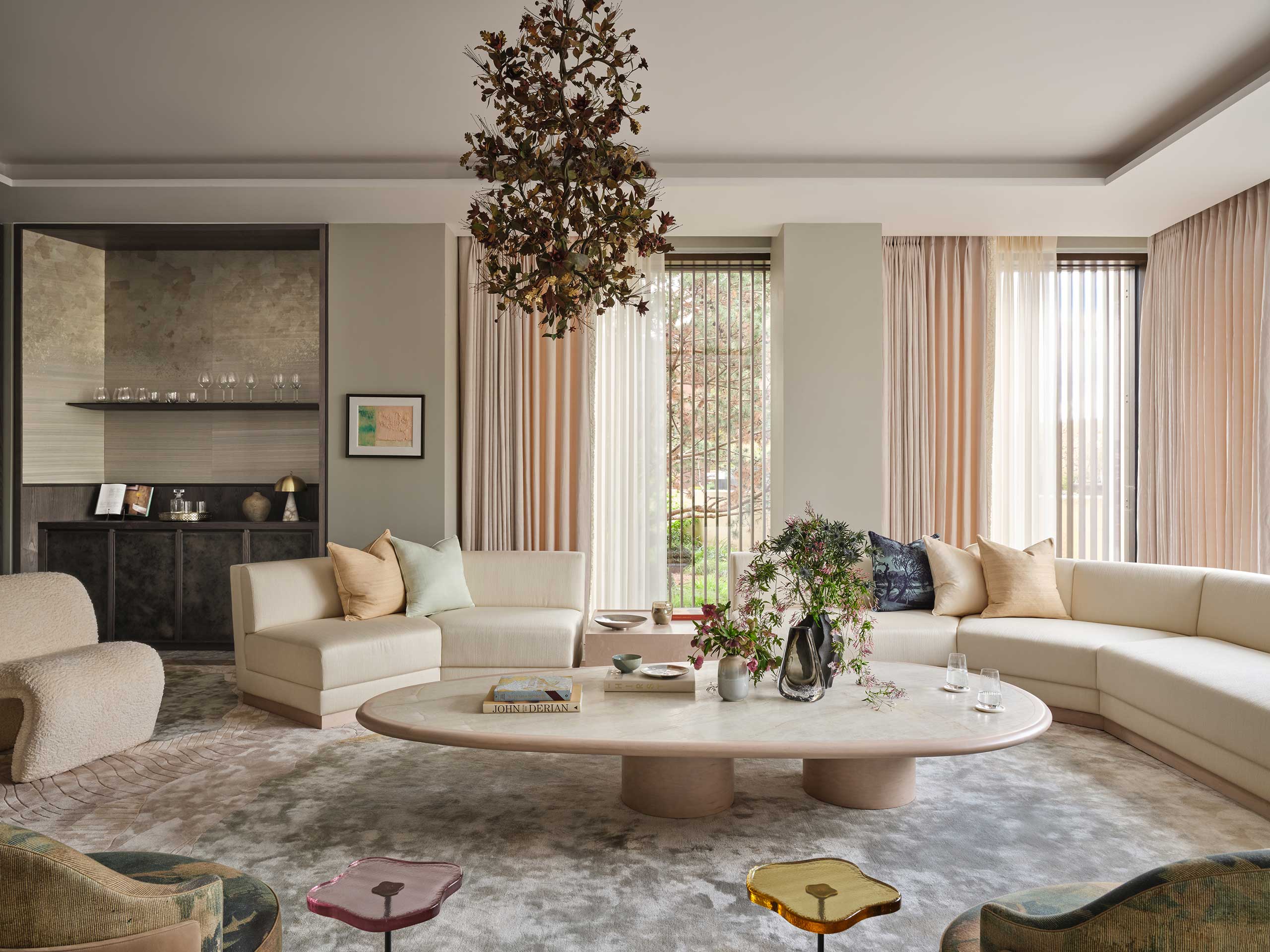 A cozy living room with beige sectional sofas, a large oval coffee table, floral centerpiece, and hanging leafy chandelier. Soft natural light filters through sheer curtains.