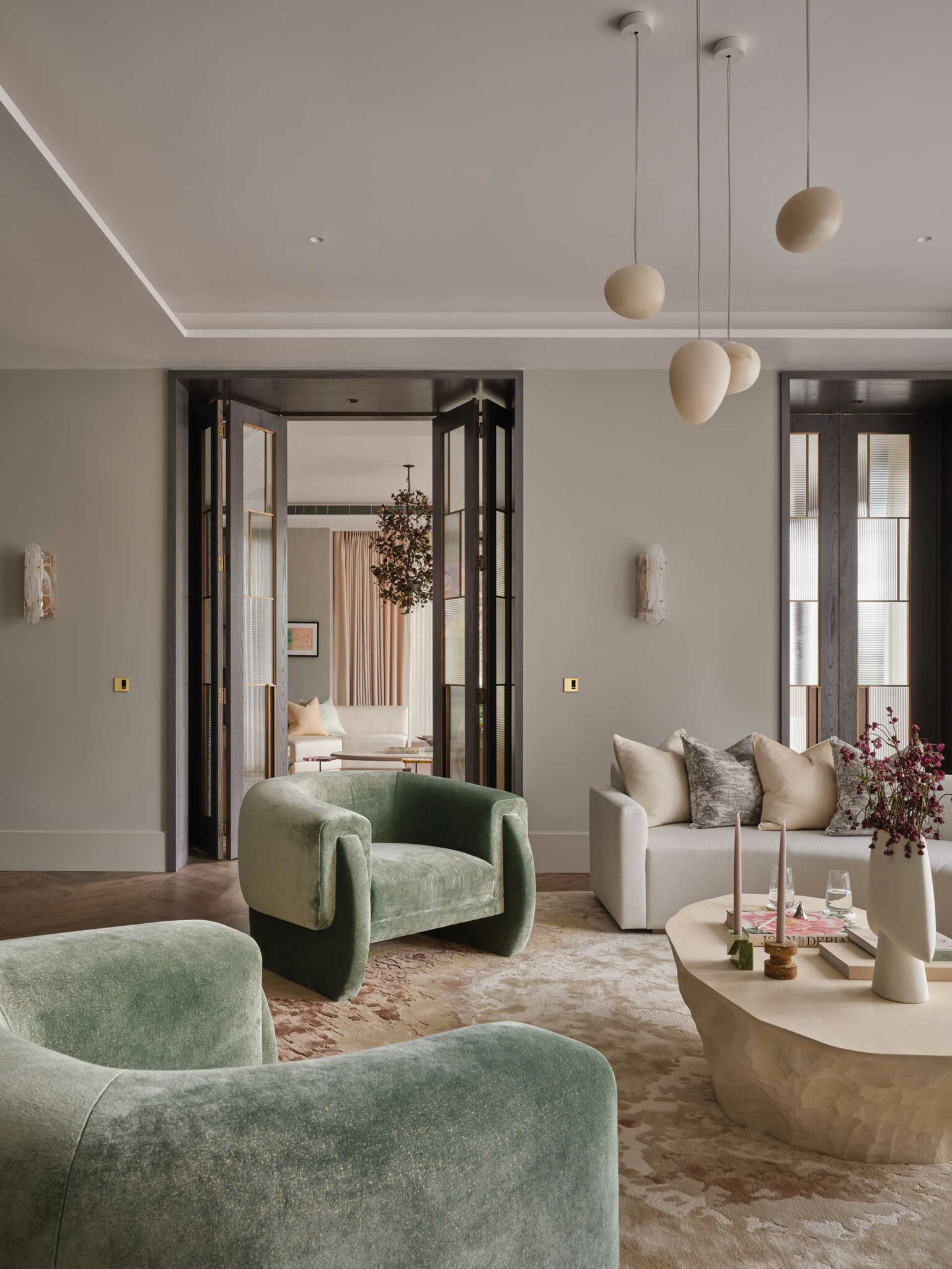 Elegant living room with sage green armchairs, beige sofa, and modern decor. Open double doors lead to another room, creating an airy, sophisticated feel.