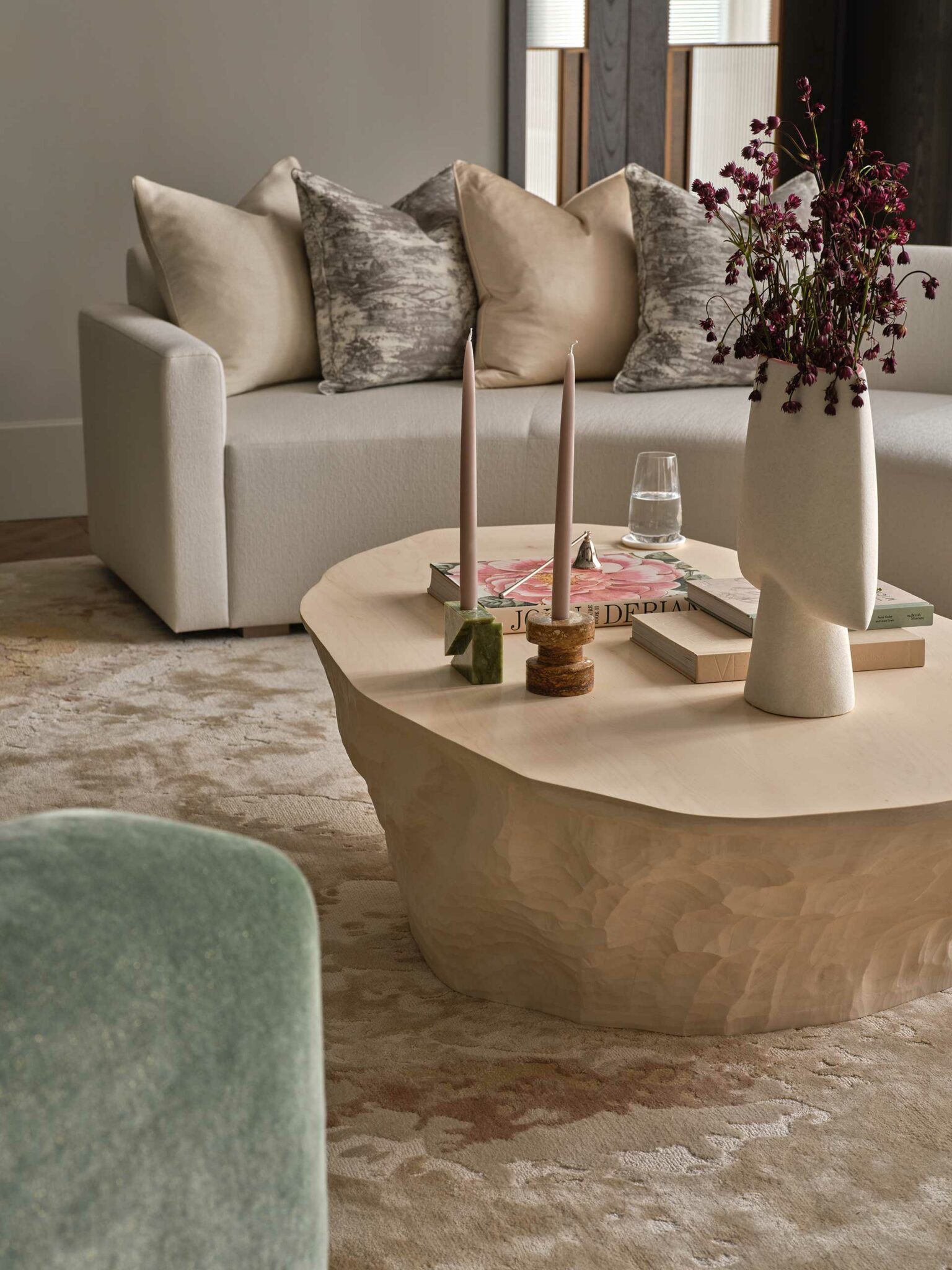 Minimalist living room with a beige couch featuring assorted throw pillows. A textured coffee table holds candles, books, and a vase with red flowers.