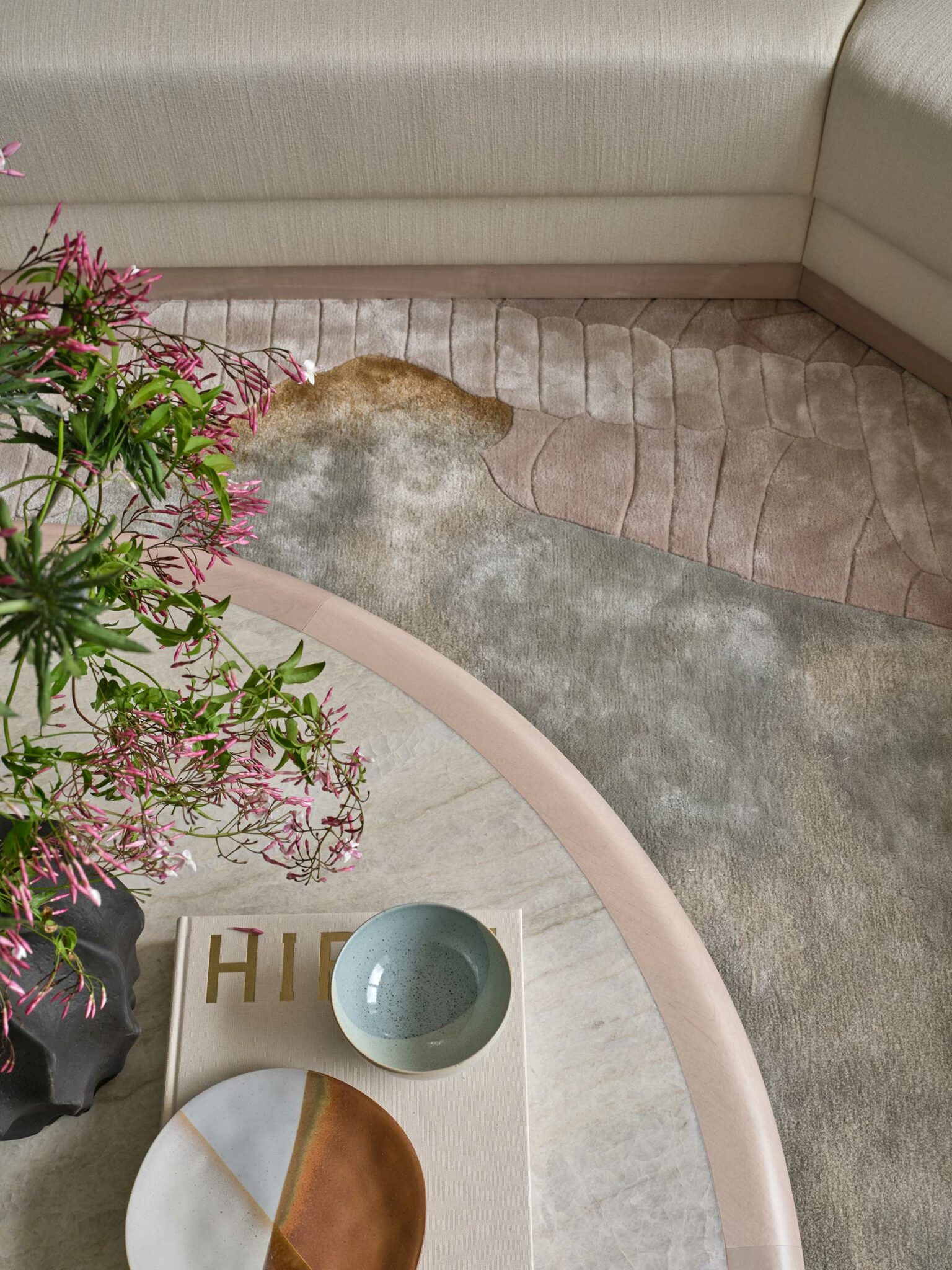 A cozy corner with a curved beige couch, abstract-patterned rug, round marble table with colorful plates, a book, and a vase of pink flowers.