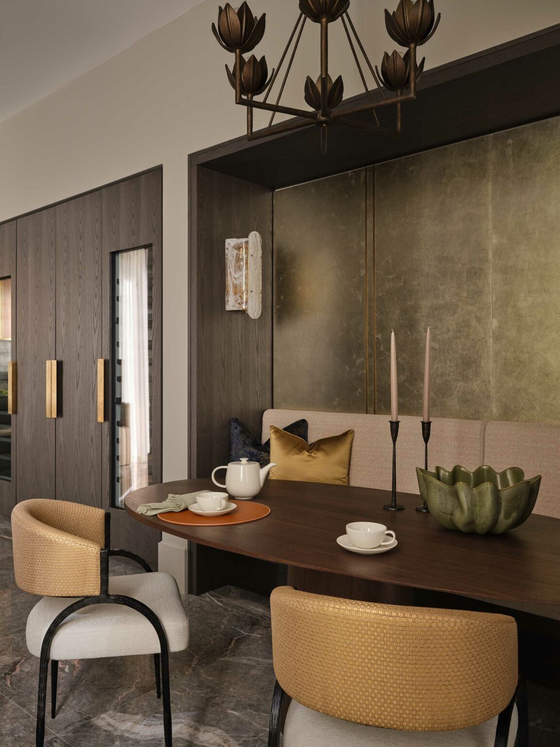 Elegant dining area with a wooden oval table, cushioned chairs, and gold accents. Candles and a teapot add warmth, under a stylish chandelier.