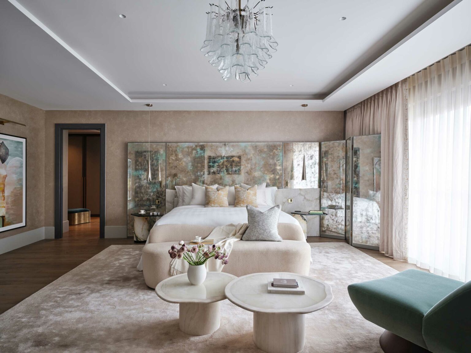 Elegant bedroom with muted tones featuring a plush bed, mirrored headboard, and delicate chandelier. Soft lighting and cozy decor evoke tranquility.
