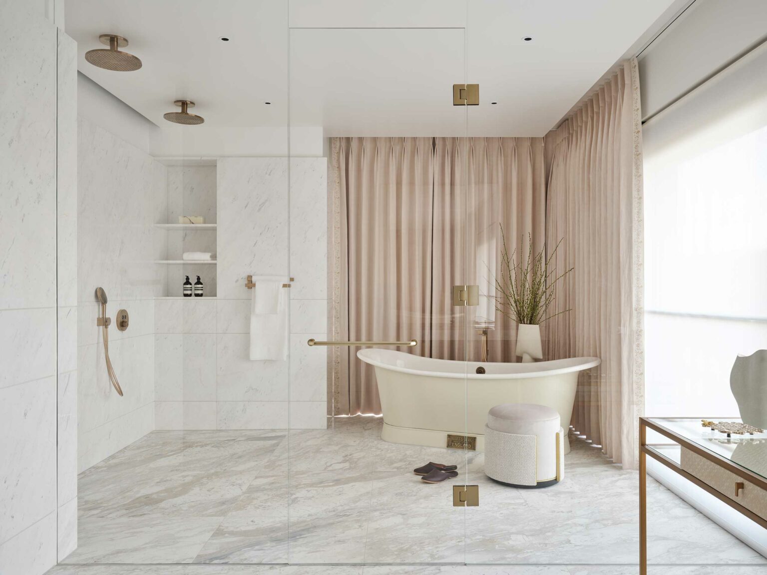 Luxurious bathroom with marble floors, beige freestanding tub, and glass shower. Soft beige curtains and a vase of reeds evoke a calm, spa-like ambiance.