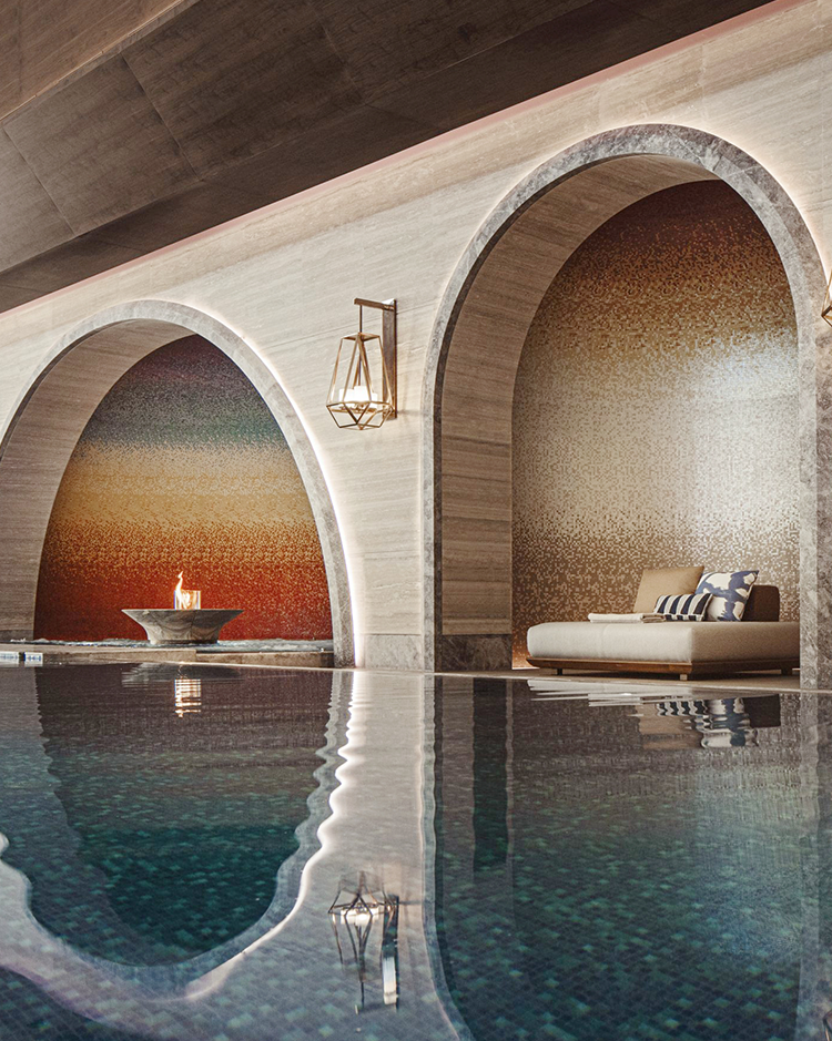 Luxurious indoor pool area with mosaic walls and elegant arches. Features a fire bowl and a cozy lounge chair with striped pillows, creating a serene ambiance.