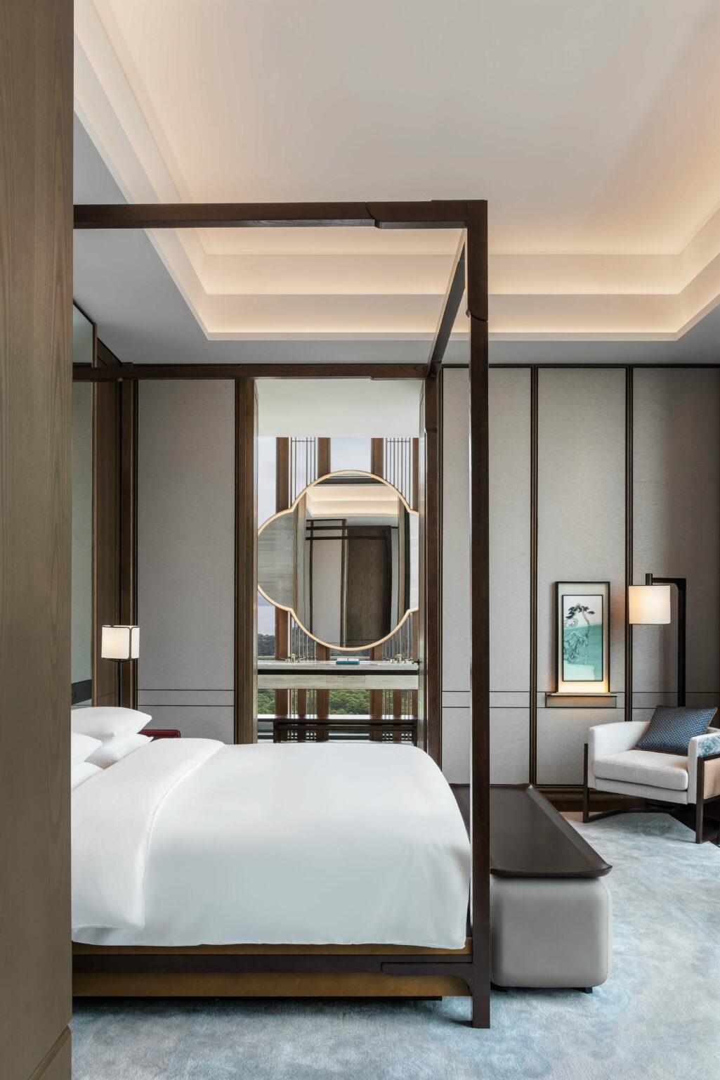 Elegant suite bedroom with a modern four-poster bed featuring crisp white linens. Soft lighting, art, and a decorative mirror create a tranquil atmosphere.