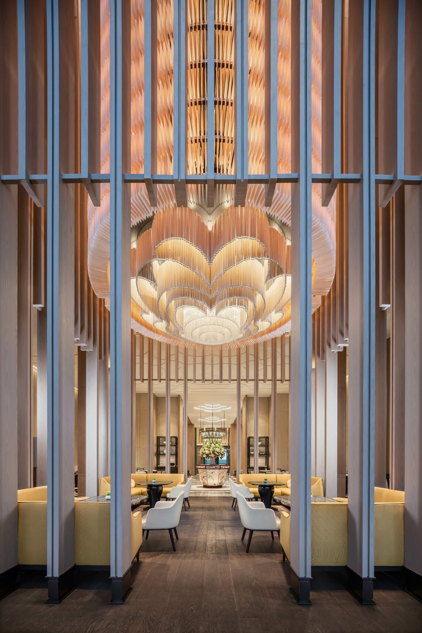 Luxurious hotel lobby with tall wooden columns and an intricate, glowing ceiling design. Yellow chairs and white armchairs create an elegant, inviting atmosphere.