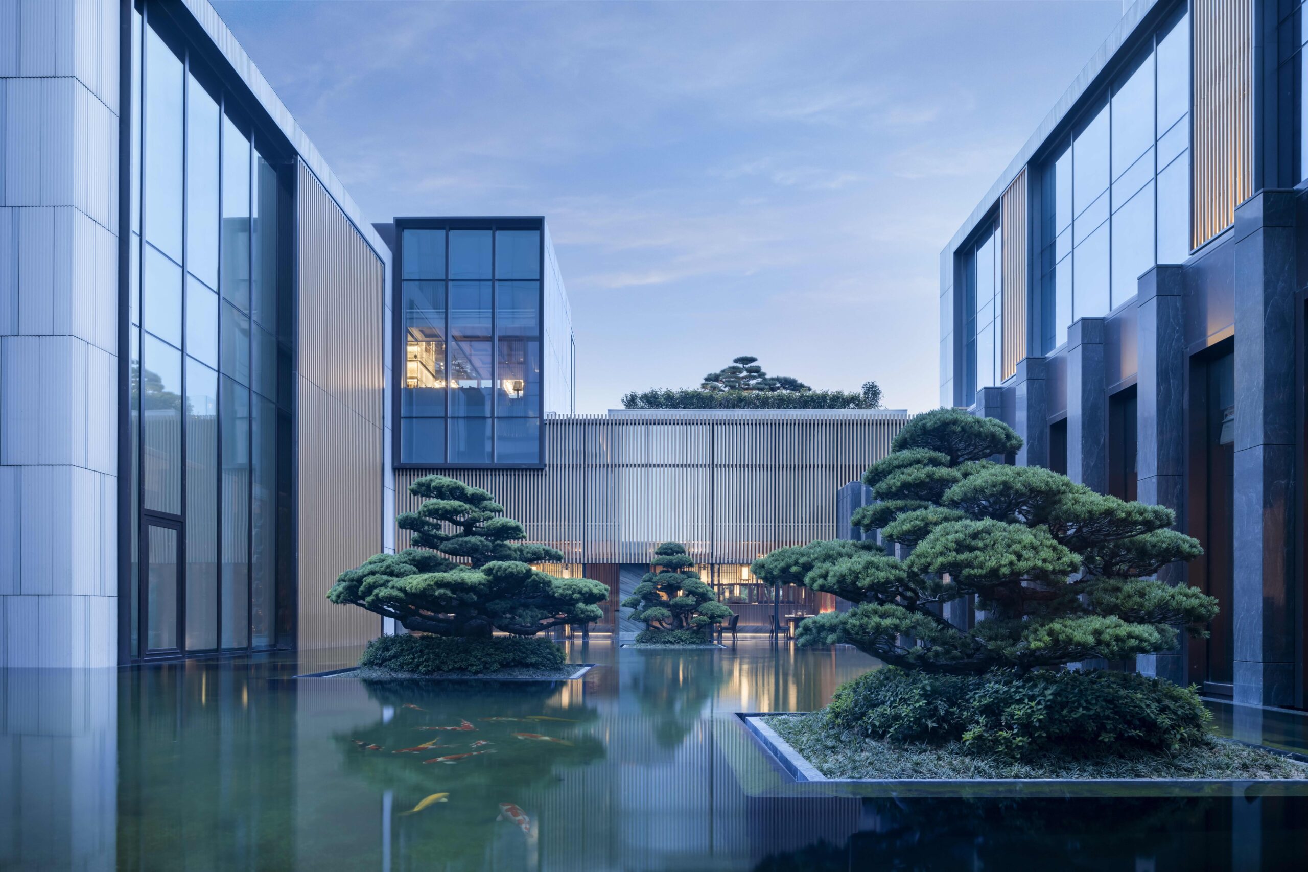 Modern hotel courtyard with a serene pond, manicured trees, and a sleek building with large glass windows under a calm blue sky, creating a tranquil atmosphere.