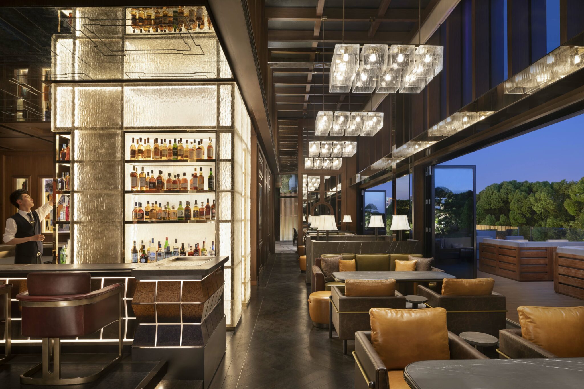 Modern hotel bar with warm lighting features a bartender selecting bottles from an illuminated shelf. Cozy seating areas and elegant chandeliers create an inviting atmosphere.