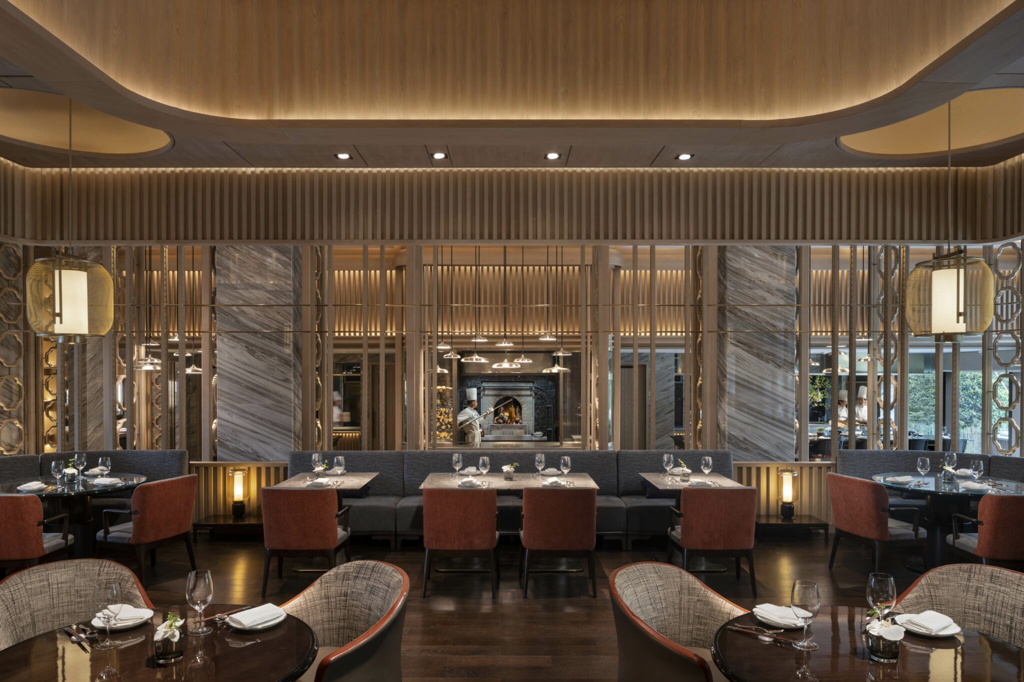 Elegant hotel restaurant interior with warm lighting, featuring neatly set tables with gray and maroon chairs, wooden accents, and a chef by a wood-fired oven.