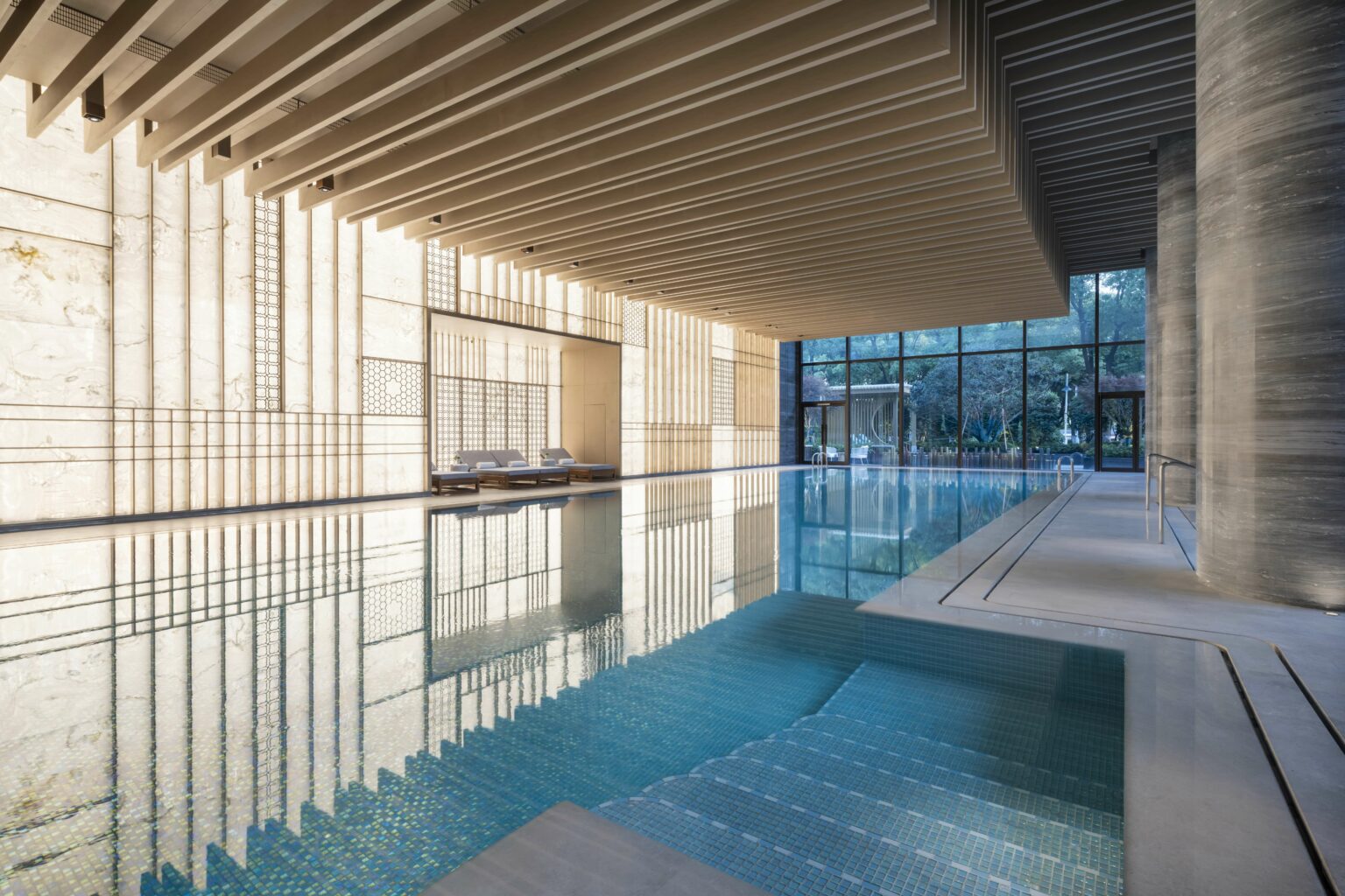 Luxurious hotel indoor swimming pool with serene blue water, elegant wood-panel ceiling, floor-to-ceiling windows, and lounge chairs, evoking tranquility.