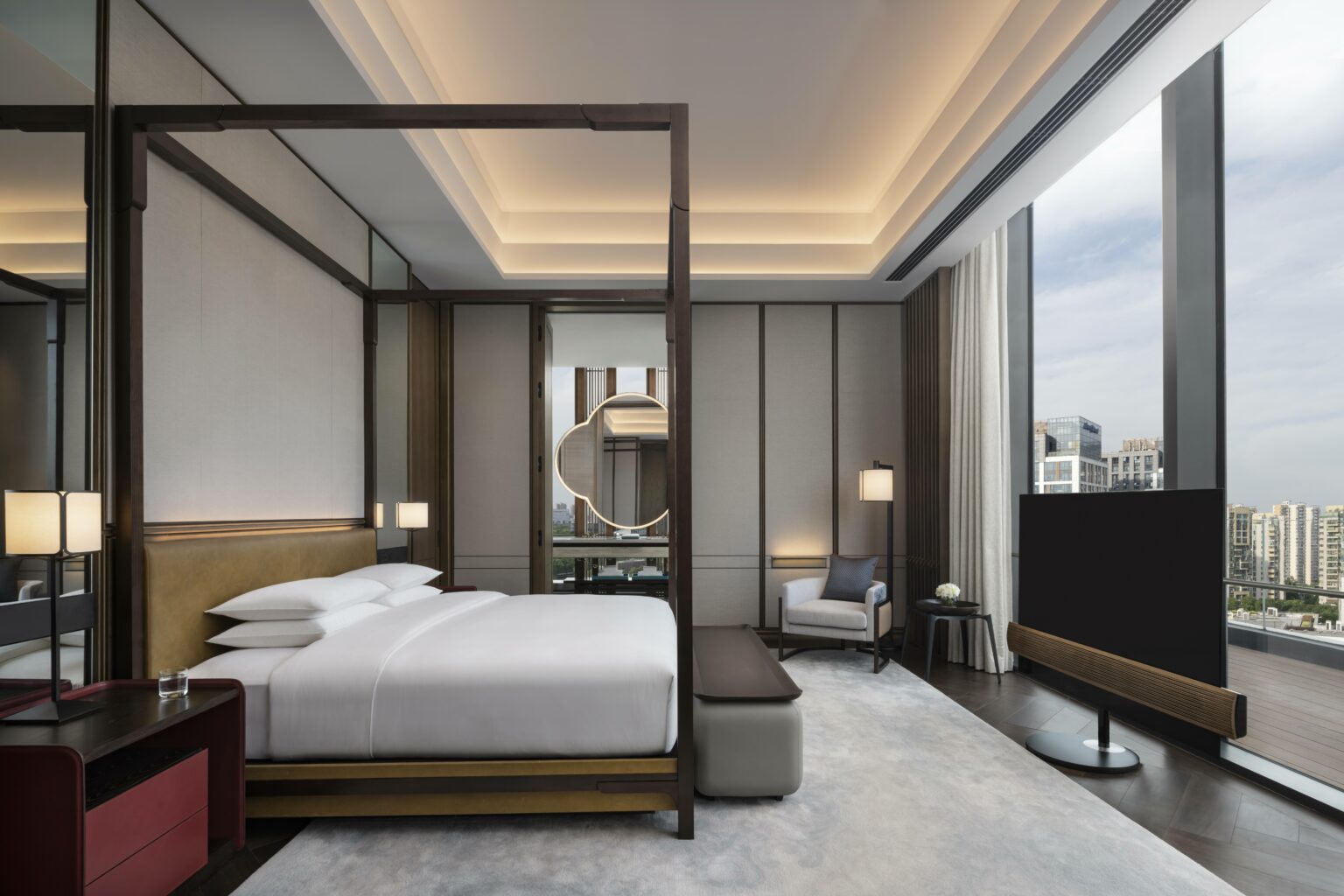 Luxurious modern suite bedroom with a large canopy bed, sleek furniture, and soft lighting. Floor-to-ceiling windows offer a cityscape view, conveying elegance and tranquility.