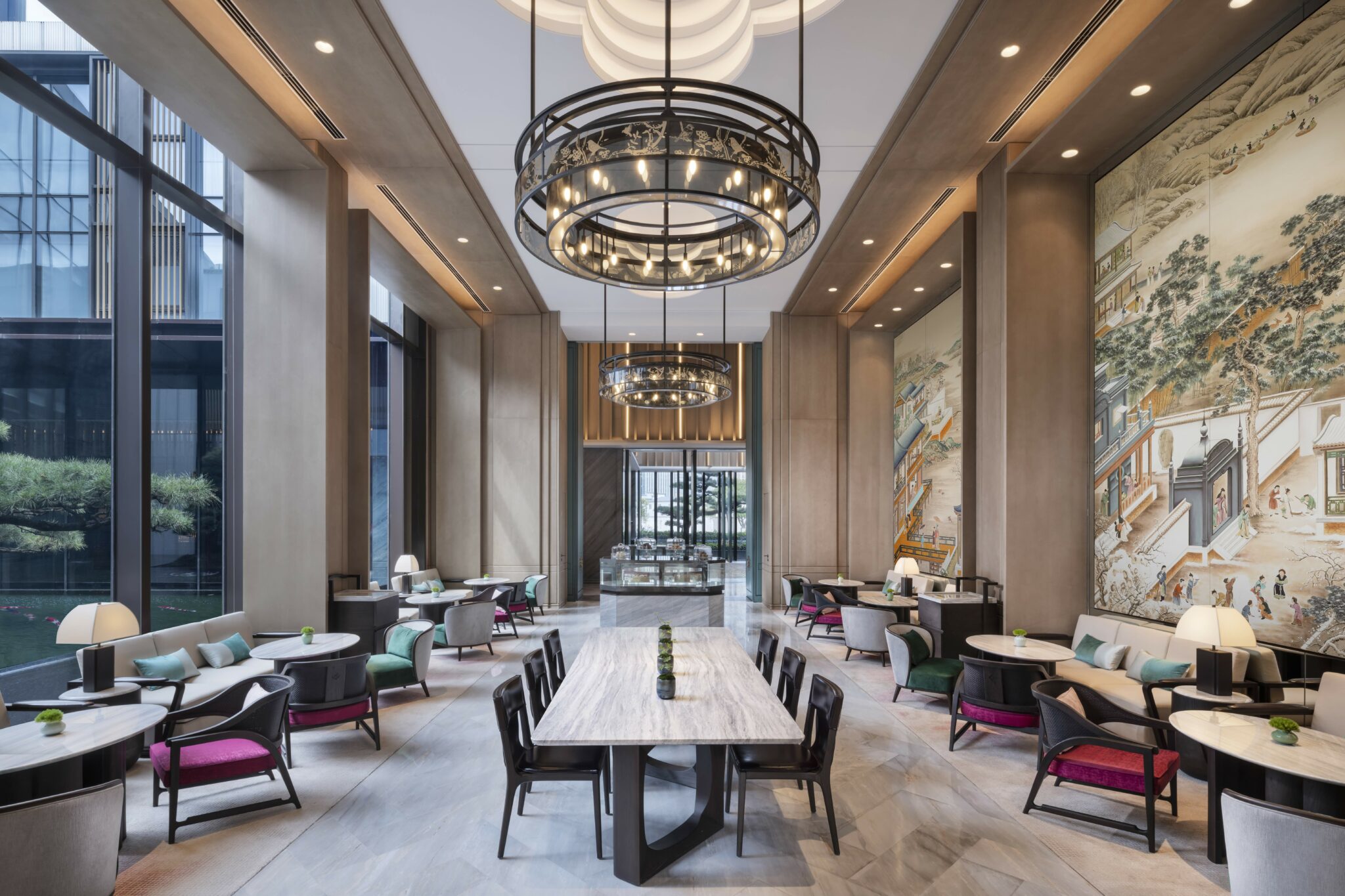 Luxurious hotel lobby with high ceilings, large chandeliers, modern seating, and a long marble table. Large windows view a garden, exuding elegance and calm.