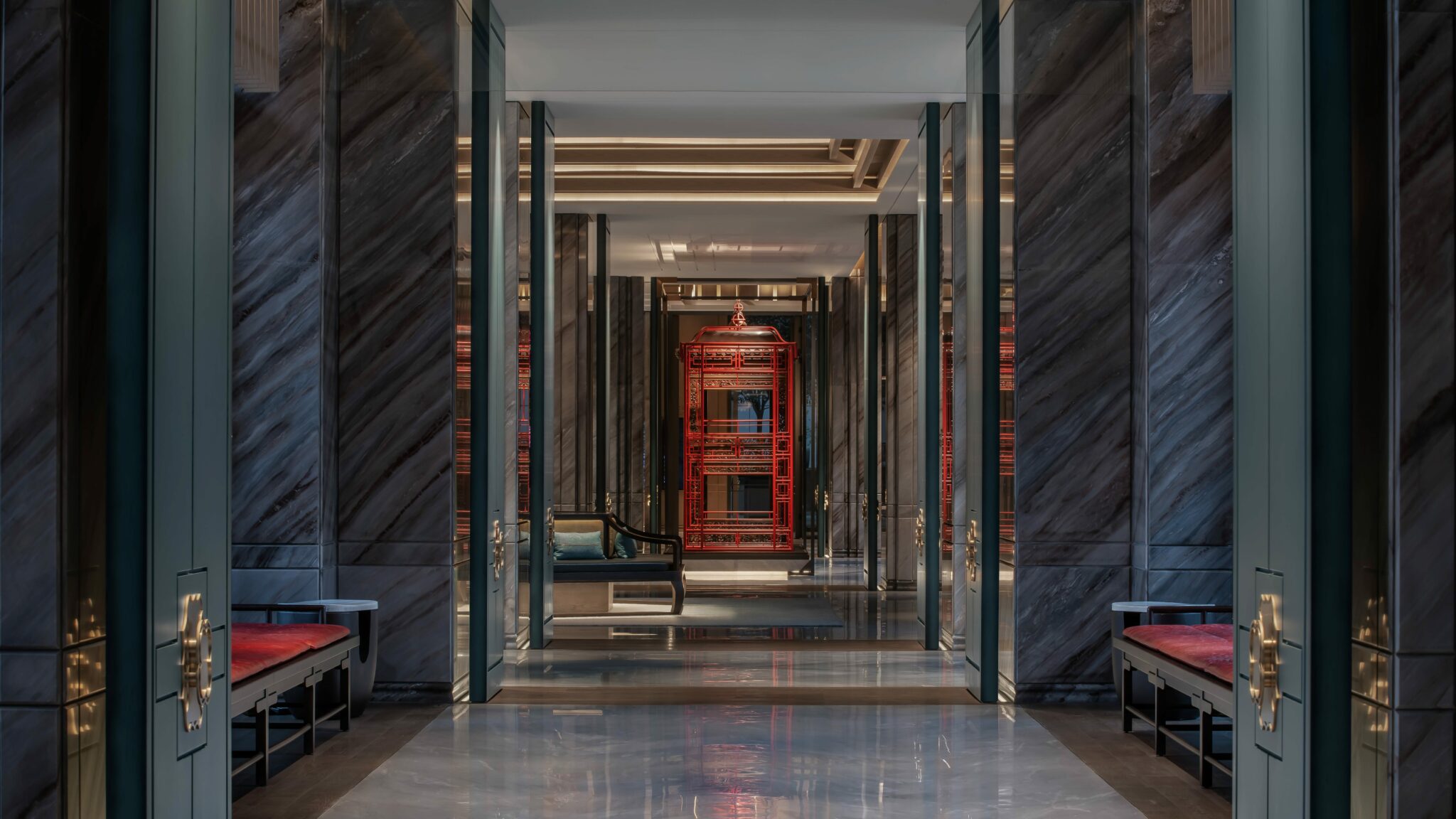 Elegant hotel hallway with marble walls and shiny floor reflecting soft lights. A vibrant red, intricate cage stands prominently at the end, framed by decorative furnishings.
