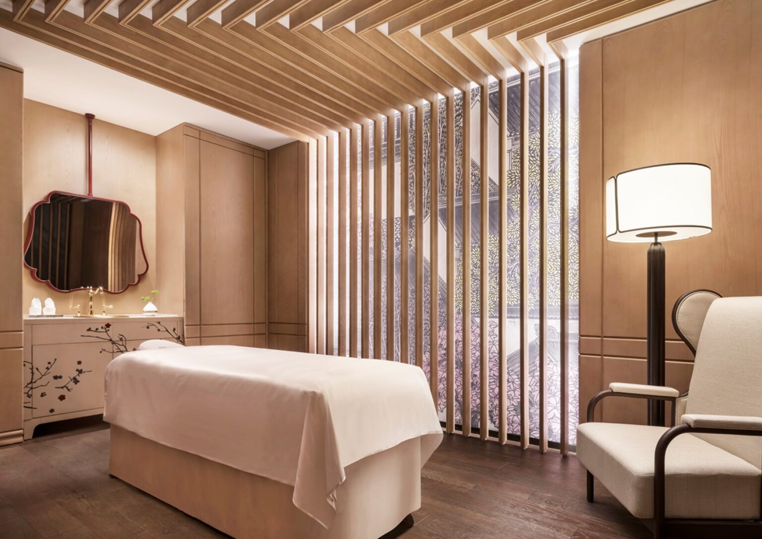 A serene hotel spa treatment room with a massage table covered in a white sheet, warm wooden paneling, decorative slats, a floral cabinet, and a soothing lamp.