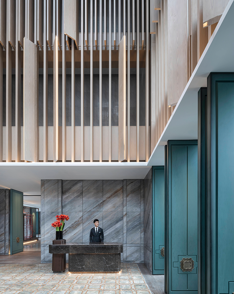 Modern hotel lobby with sleek marble walls and vertical wood slats. A suited receptionist stands behind a dark marble desk, beside vibrant red flowers.