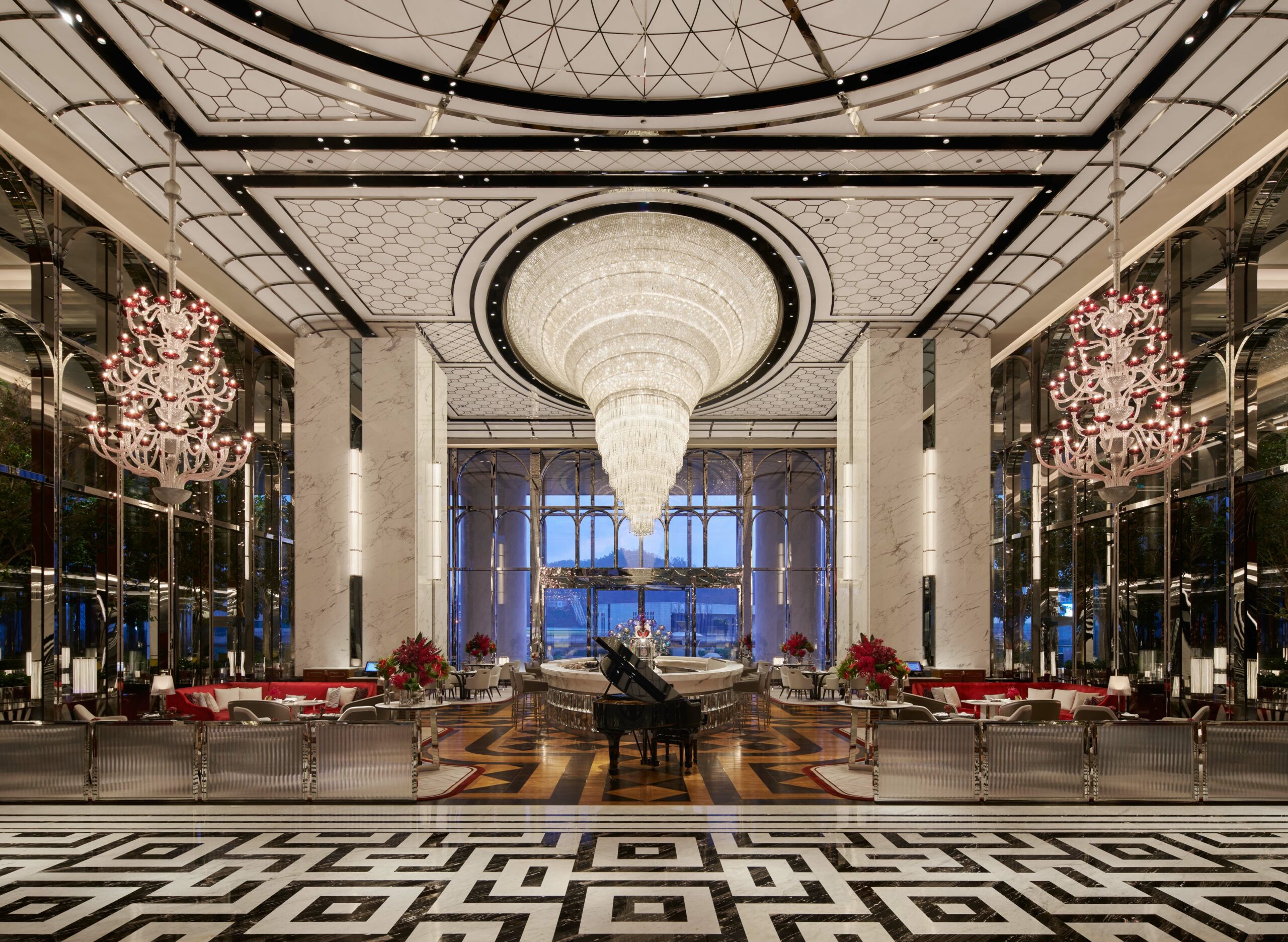 Luxurious hotel lobby with a grand spiral chandelier, elegant seating, and a black piano. Marble columns and geometric floor enhance the opulent ambiance.