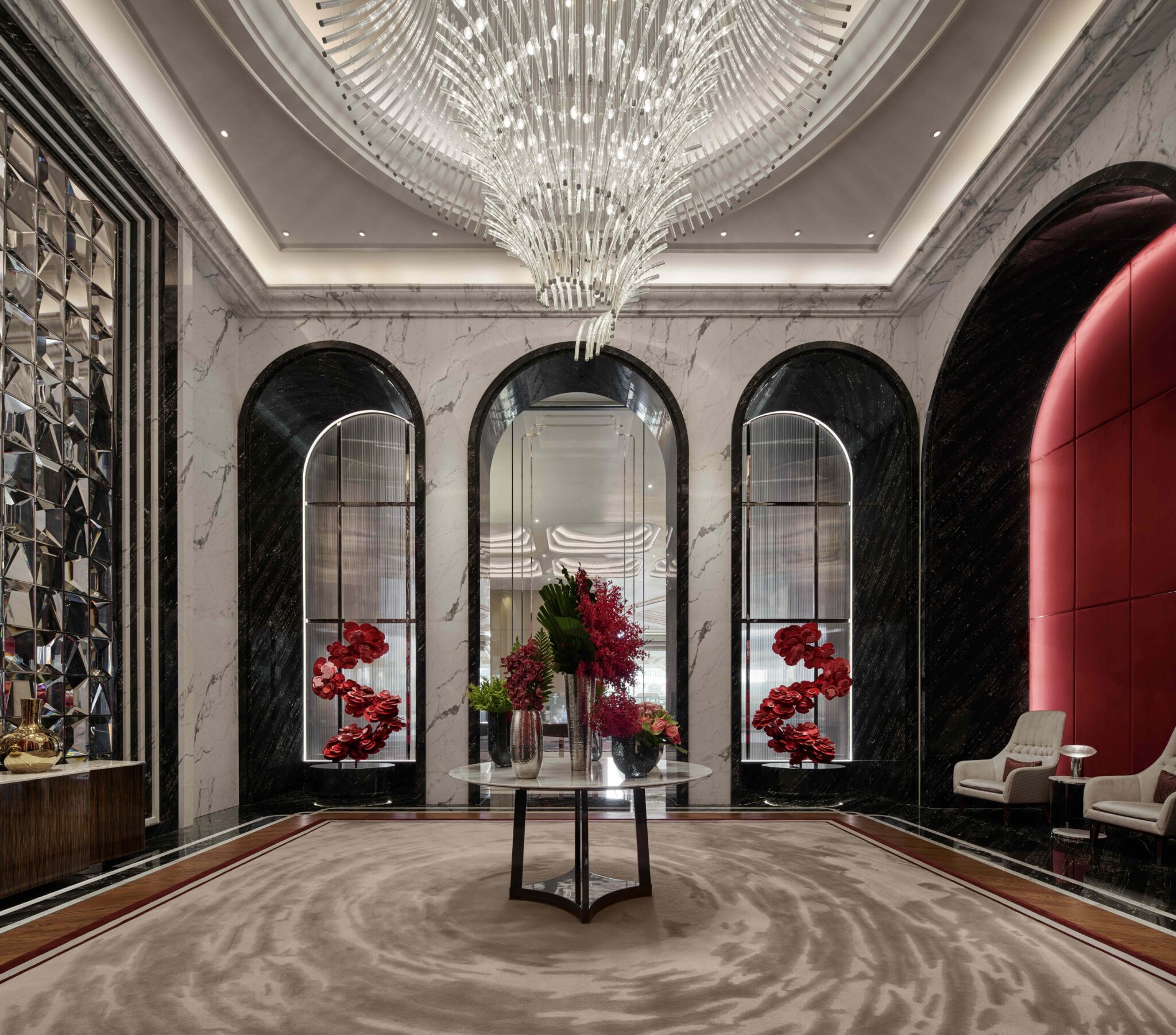 Luxurious hotel lobby with grand chandelier, marble walls, and arched mirrors. A round table holds vibrant red-flowered vases, exuding elegance.