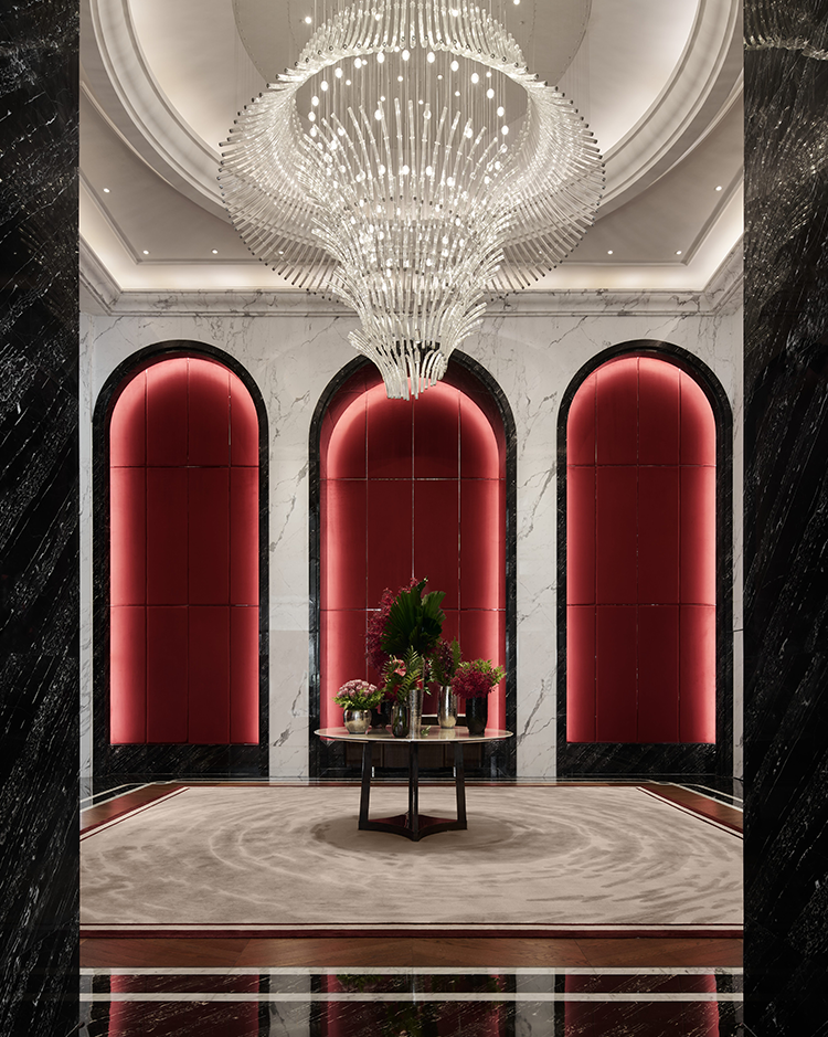 A luxurious hotel lobby featuring a grand chandelier, elegant arched design elements, and rich red accents, complemented by a central table adorned with flowers, creating a sophisticated and welcoming ambiance.