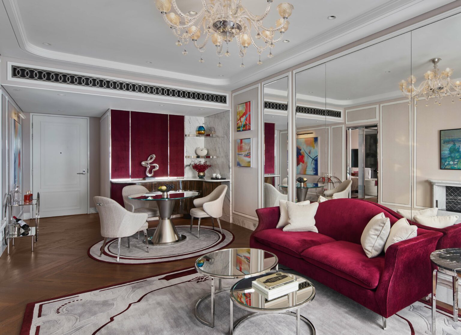 Luxurious suite living room with a deep red velvet sofa, glass tables, and beige chairs. Elegant chandeliers and mirrored walls enhance the sophisticated decor.