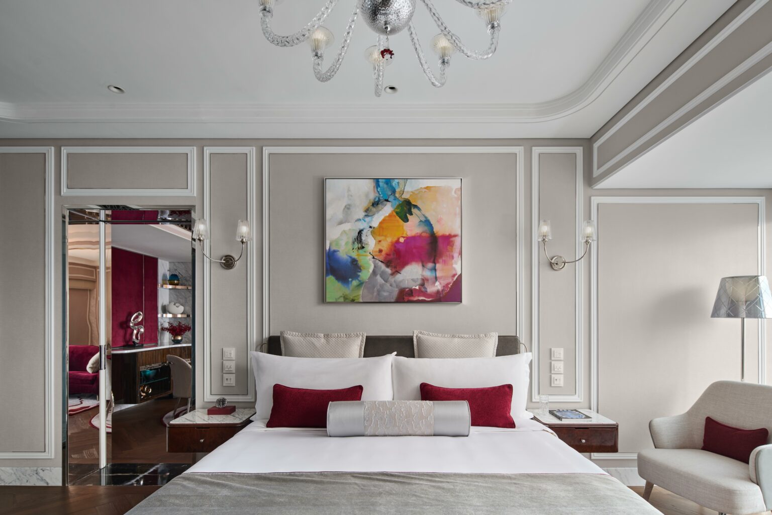 Luxurious suite bedroom with a modern aesthetic, featuring a large abstract painting above a king-sized bed with red and gray accents, soft lighting, and plush furnishings.