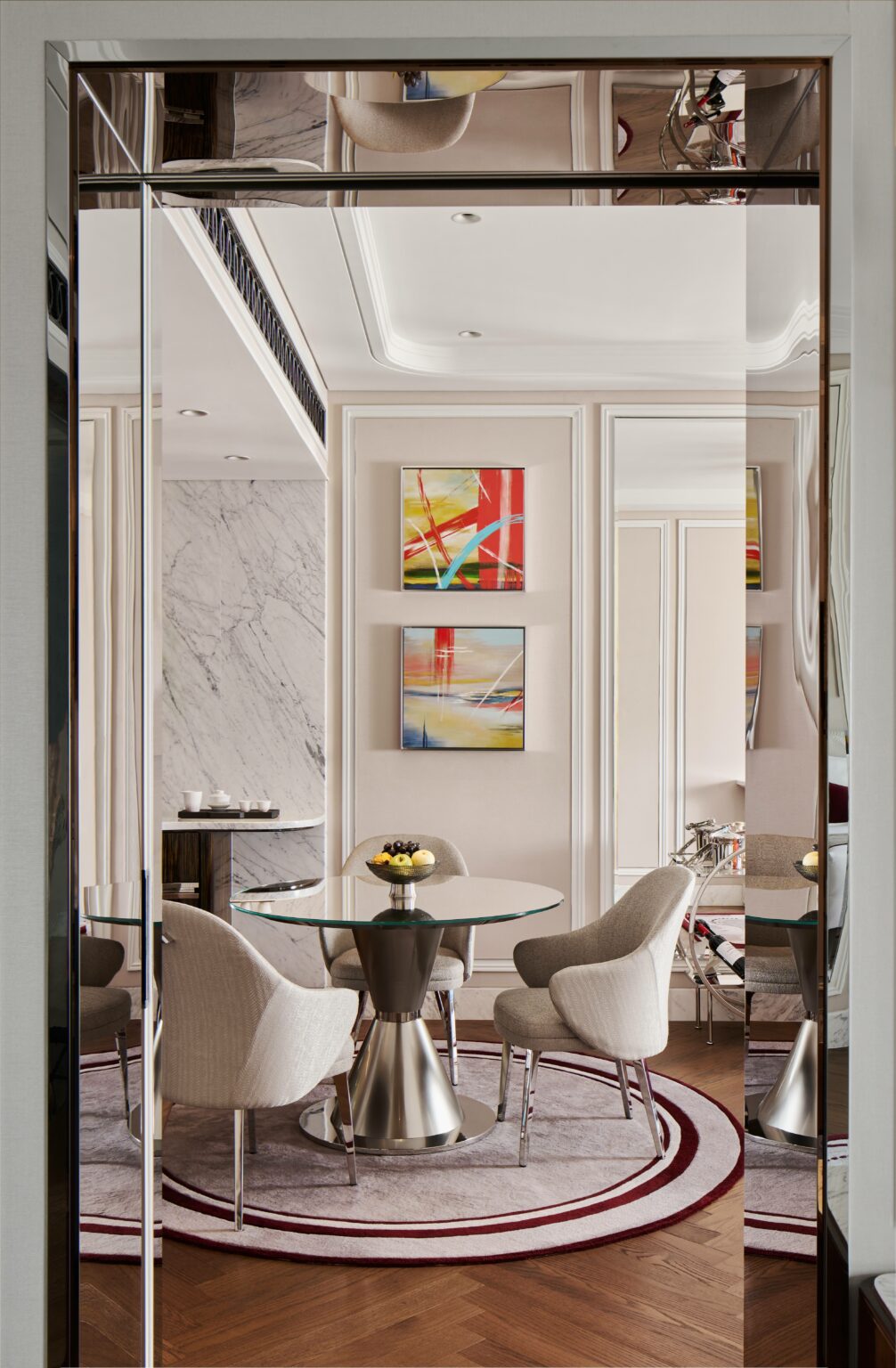 Modern suite dining room area with glass table, silver base, and four gray chairs on a round rug. Abstract art on walls, marble accent, and a warm ambiance.