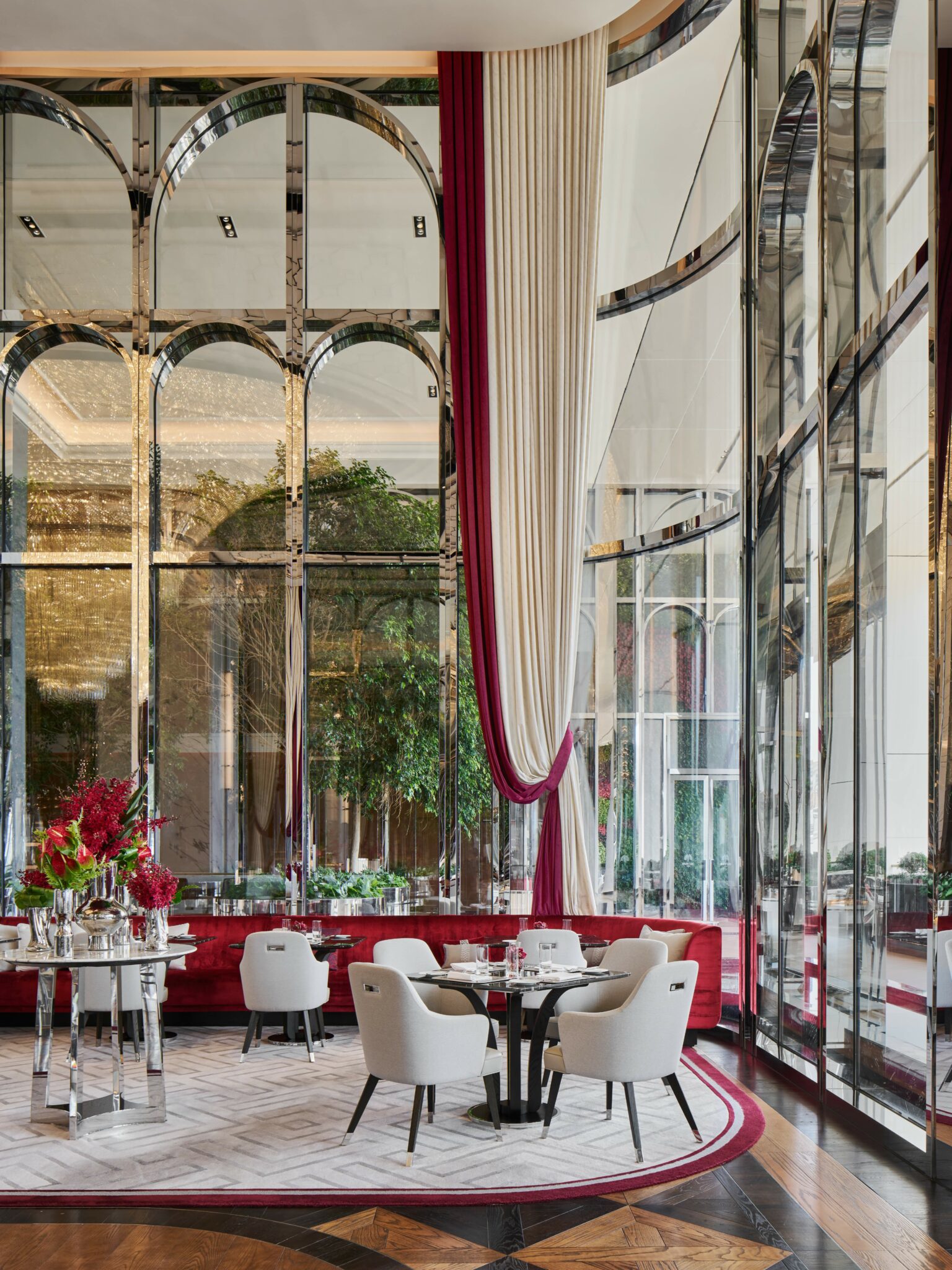 Elegant hotel dining area with glass walls, arched mirrors, and lush plants. Beige chairs around a round table, with red accents and draped curtains.