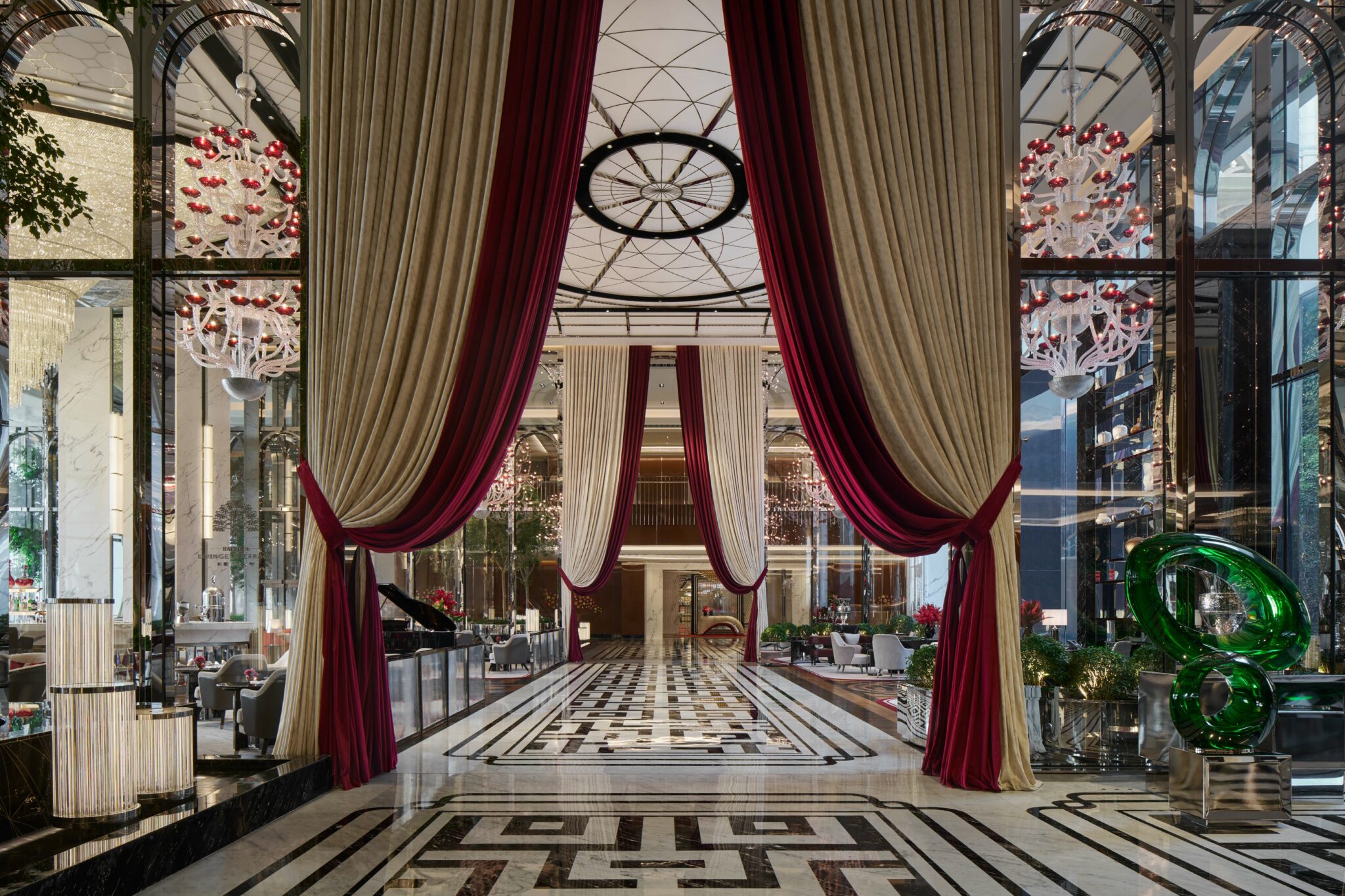 Luxurious hotel lobby with grand red and cream drapes, elaborate chandeliers, and a black-and-white patterned floor. Elegant and welcoming atmosphere.