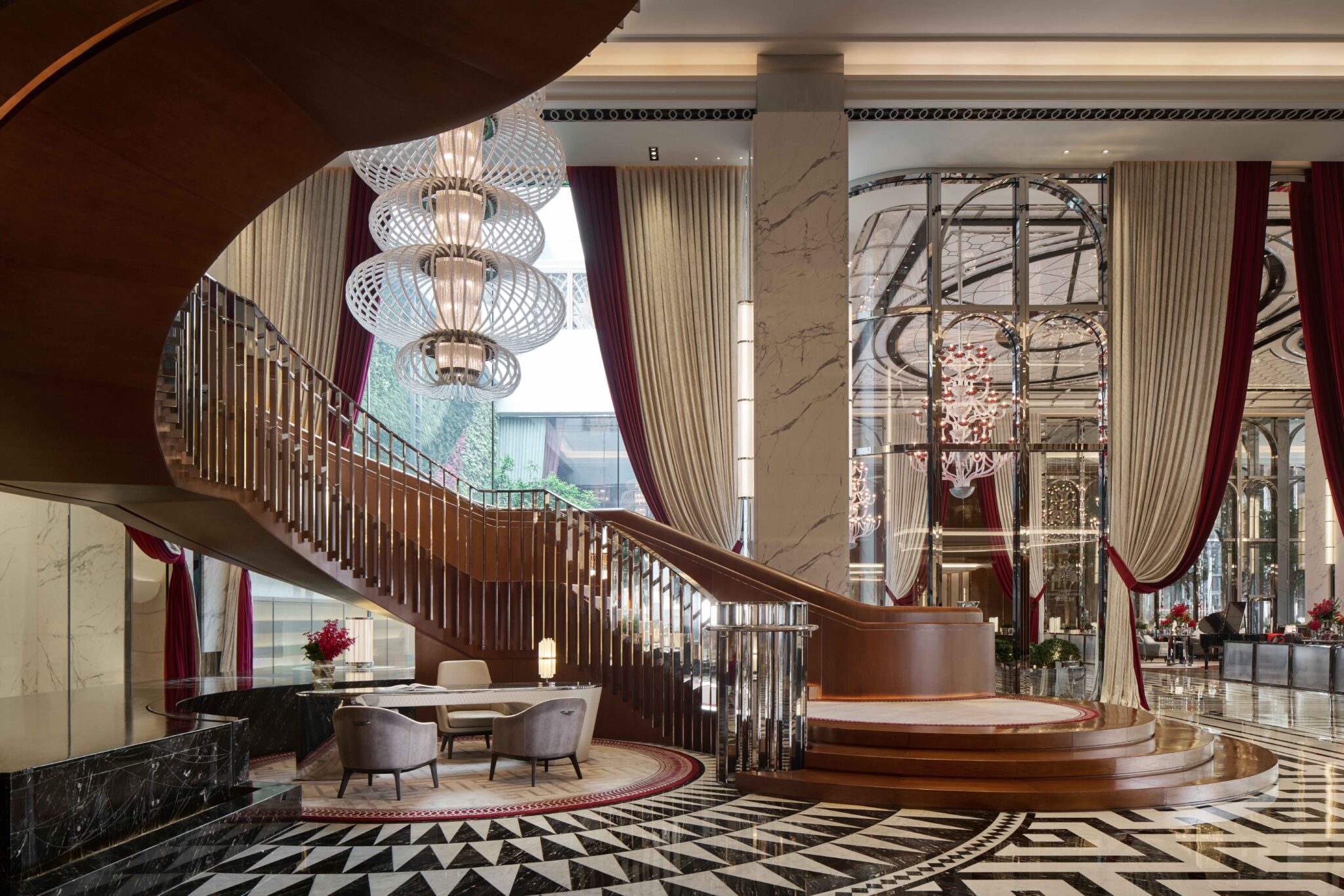 Luxurious hotel lobby with a sweeping wooden staircase, circular chandelier, marble columns, red and cream drapes, and elegant seating area.