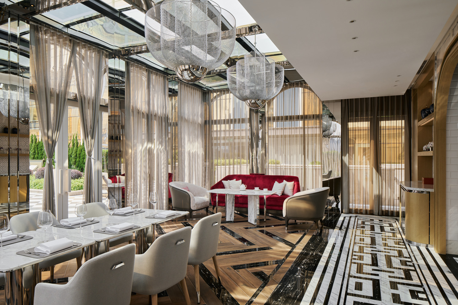 Elegant hotel dining area with glass walls, arched mirrors, and lush plants. Beige chairs around a round table, with red accents and draped curtains.