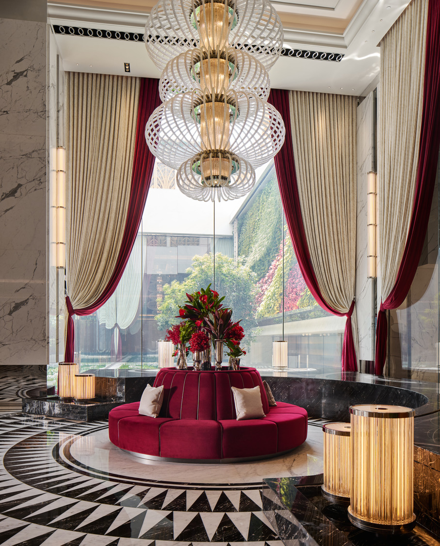Luxurious hotel lobby with grand red and cream drapes, elaborate chandeliers, and a black-and-white patterned floor. Elegant and welcoming atmosphere.