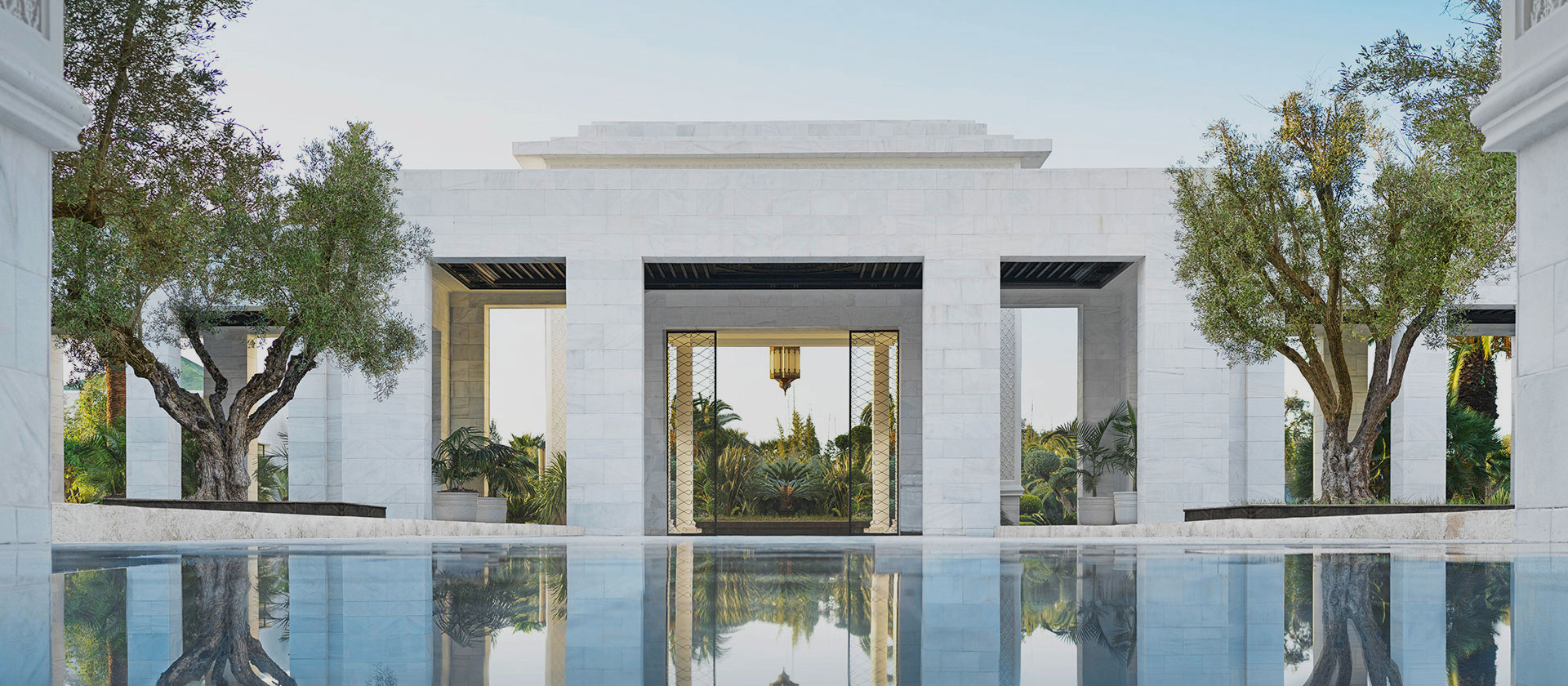 Elegant white marble structure with open arches, framed by lush trees. A serene reflecting pool mirrors the scene, evoking tranquility and luxury.