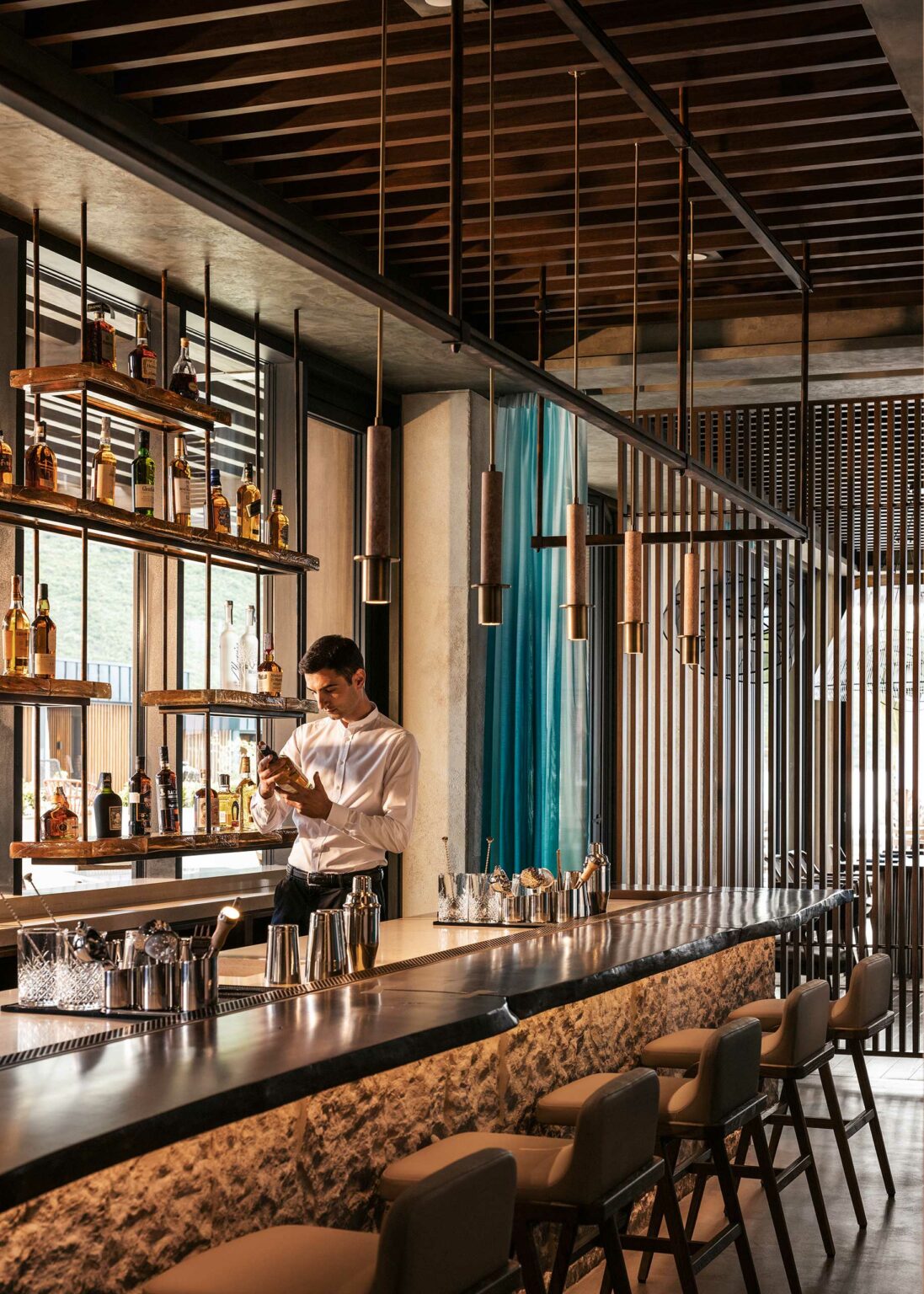 Bartender in a sleek, modern bar shakes a cocktail. Shelves with liquor bottles are behind him, and pendant lights hang from the ceiling, creating a warm ambiance.