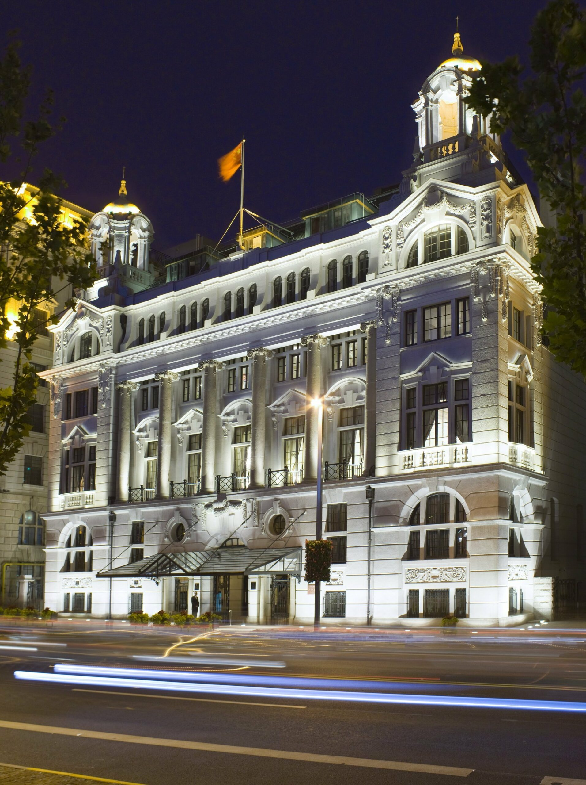 Waldorf Astoria Shanghai's exterior. Illuminated at night, a grand neoclassical building with ornate arches and columns is adorned with a flag on top.
