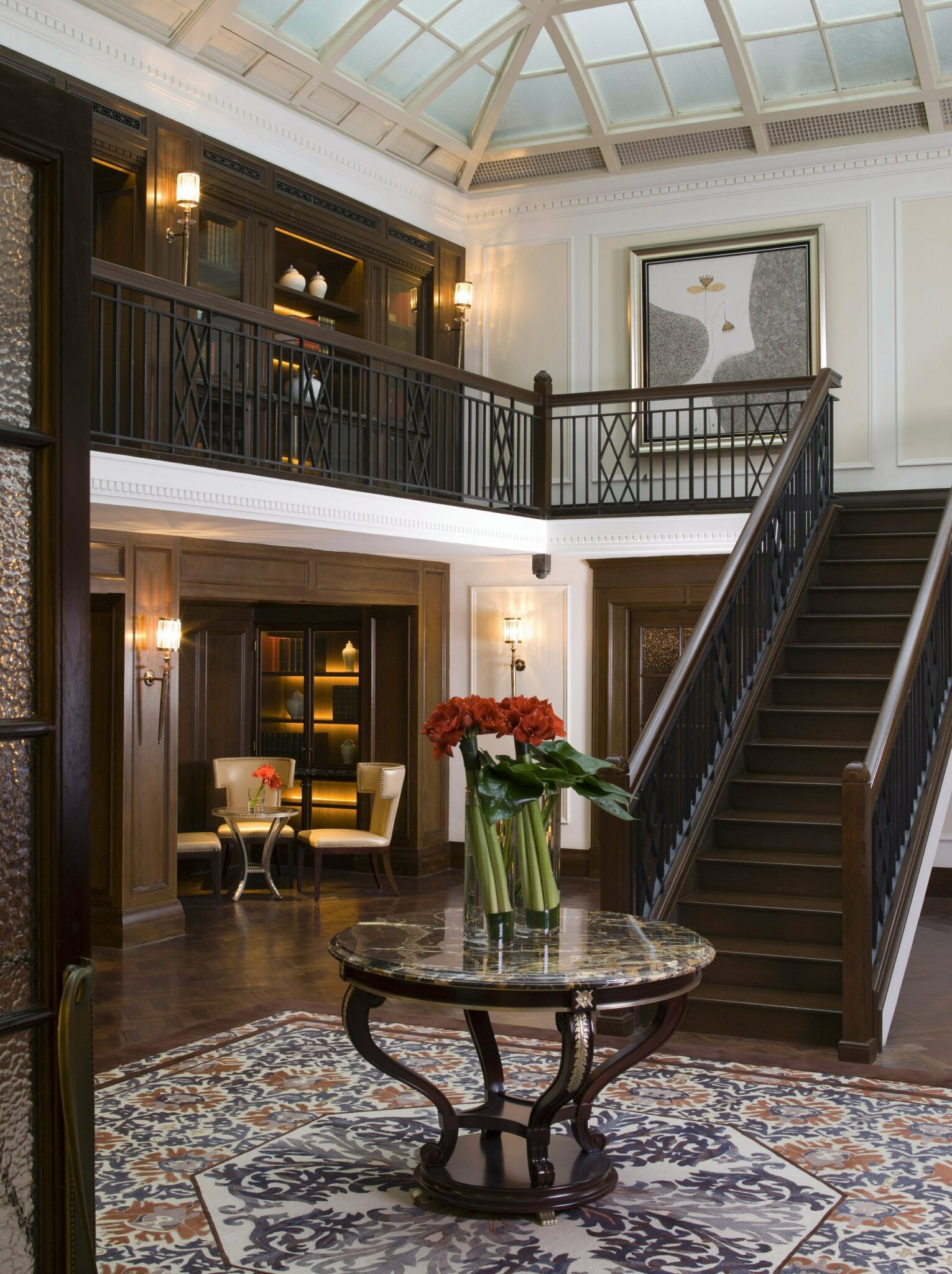 Elegant library foyer with wood paneling, a round table with red flowers, and a staircase leading to a mezzanine. Warm lighting creates a cozy ambiance.