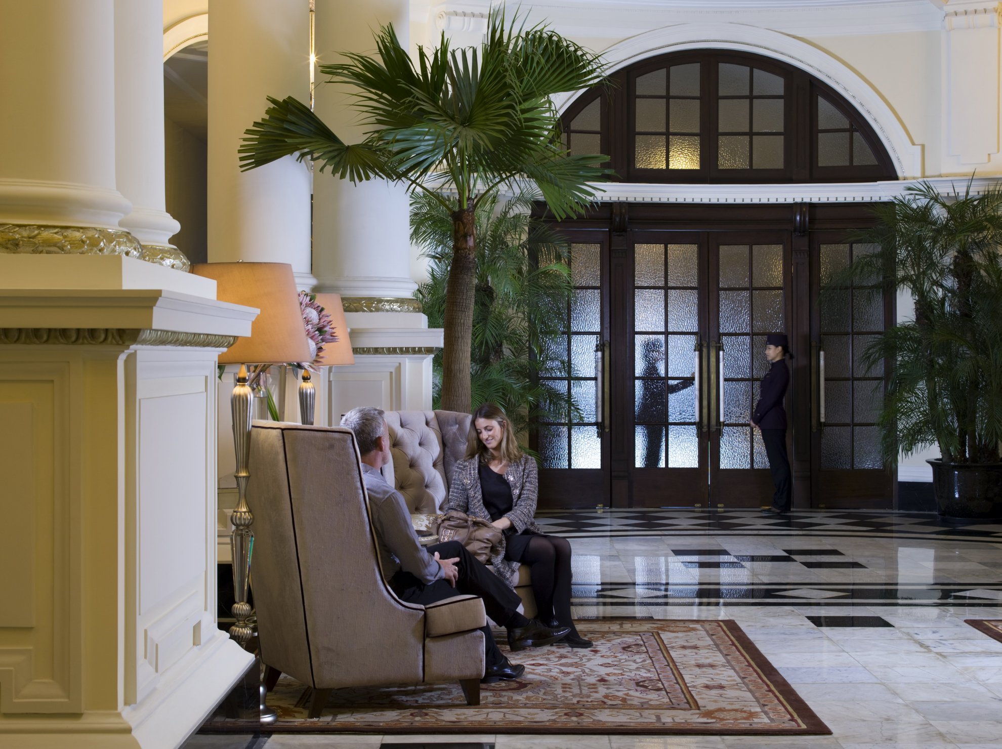 A couple sits in a grand hotel lobby, engaged in conversation on plush chairs. A doorman stands by large wooden doors. Tall palms and elegant decor set a sophisticated tone.