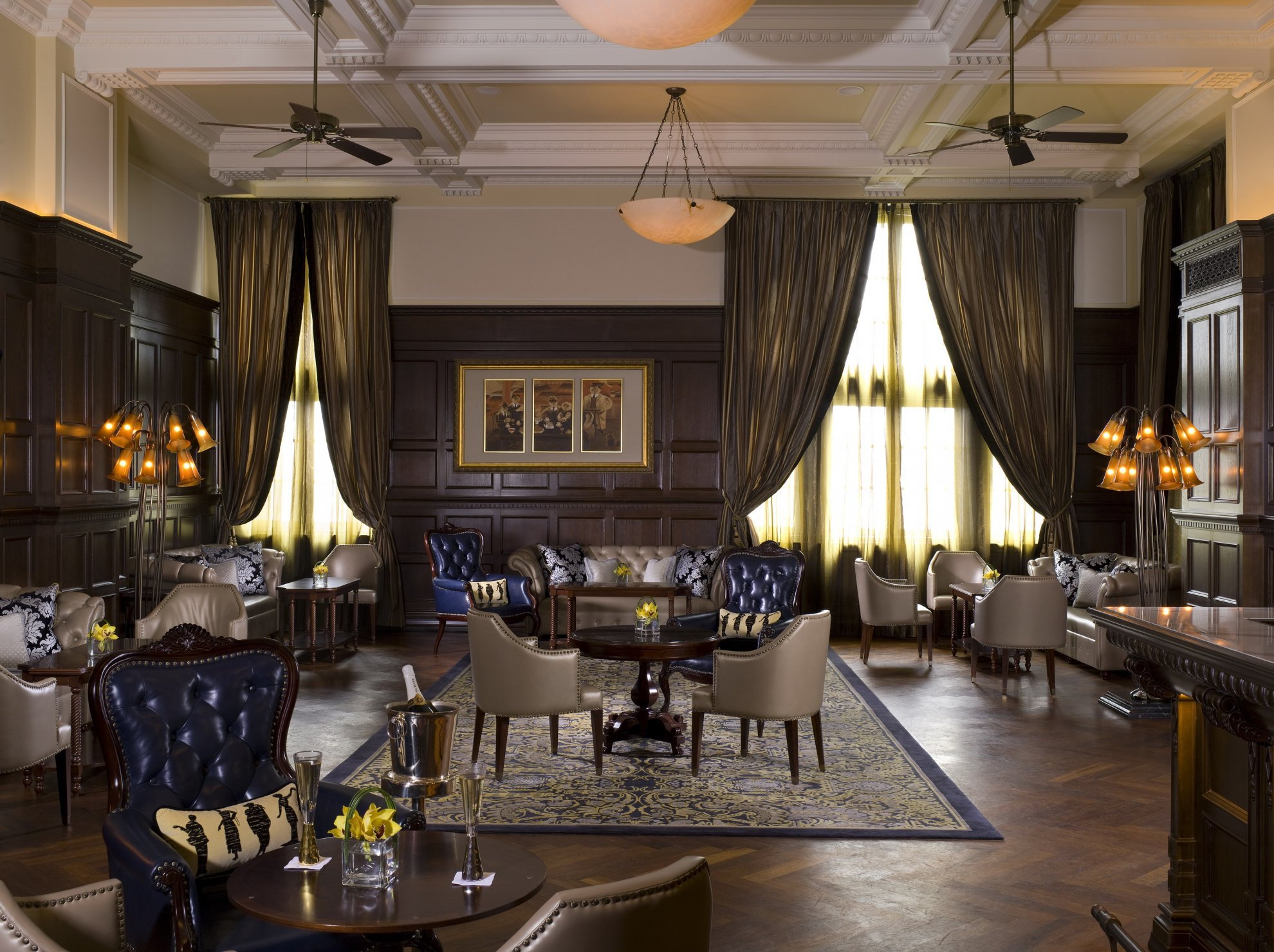 Elegant hotel lounge with dark wood panels and ambient lighting. Leather armchairs, sofas, and tables are arranged on a patterned carpet. Plush drapes frame large windows.