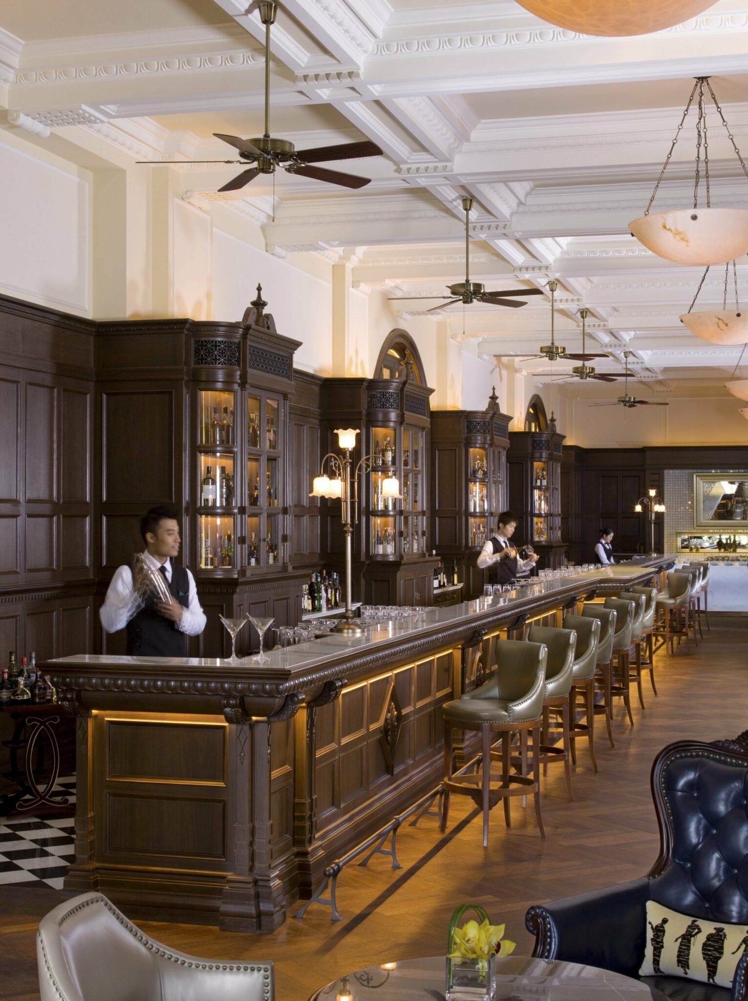 A luxurious bar with dark wood cabinetry, elegant lighting, and plush seating, featuring bartenders preparing drinks behind the polished counter.
